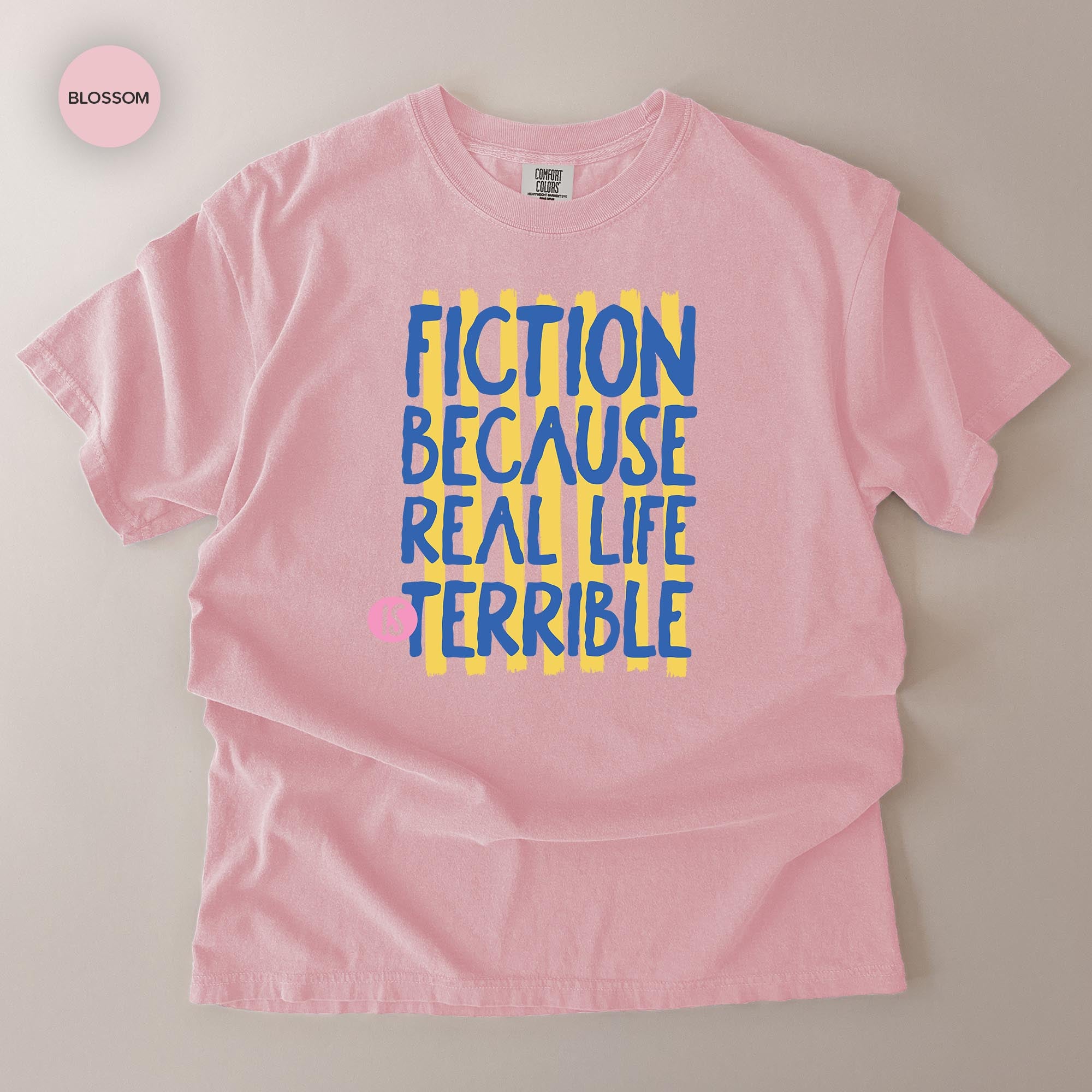 A pink t-shirt with a printed message that reads "FICTION BECAUSE REAL LIFE TERRIBLE" in blue and yellow text.