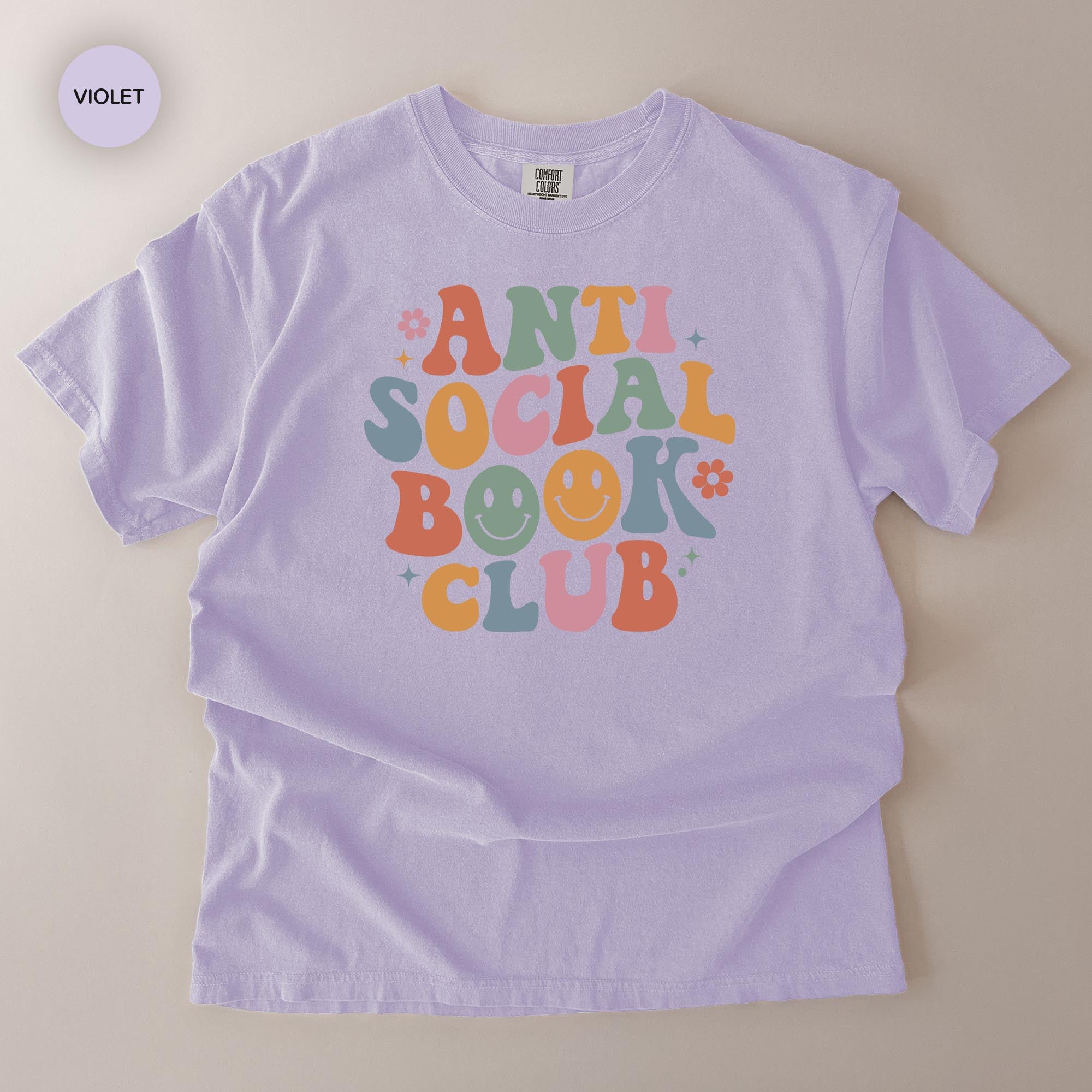 A purple t-shirt with a colorful graphic design featuring the text "ANTI SOCIAL BOOK CLUB" in a retro style.