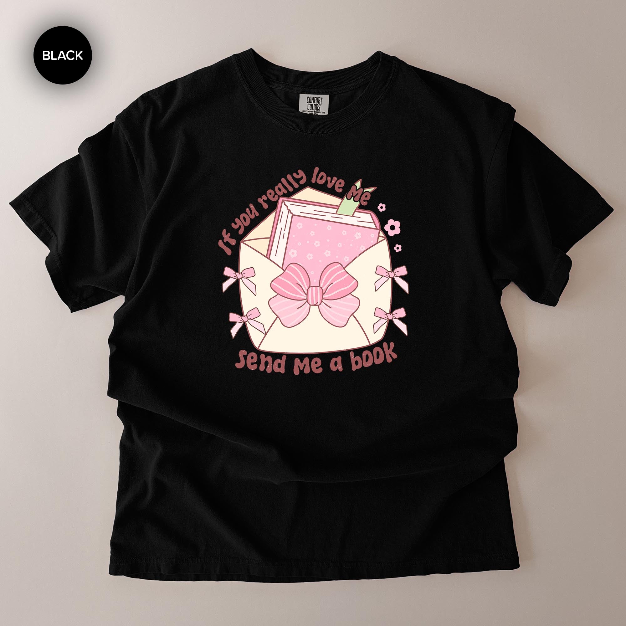 A black t-shirt with a graphic design featuring a pink bow, a book, and the text "If you really love me, send me a book".