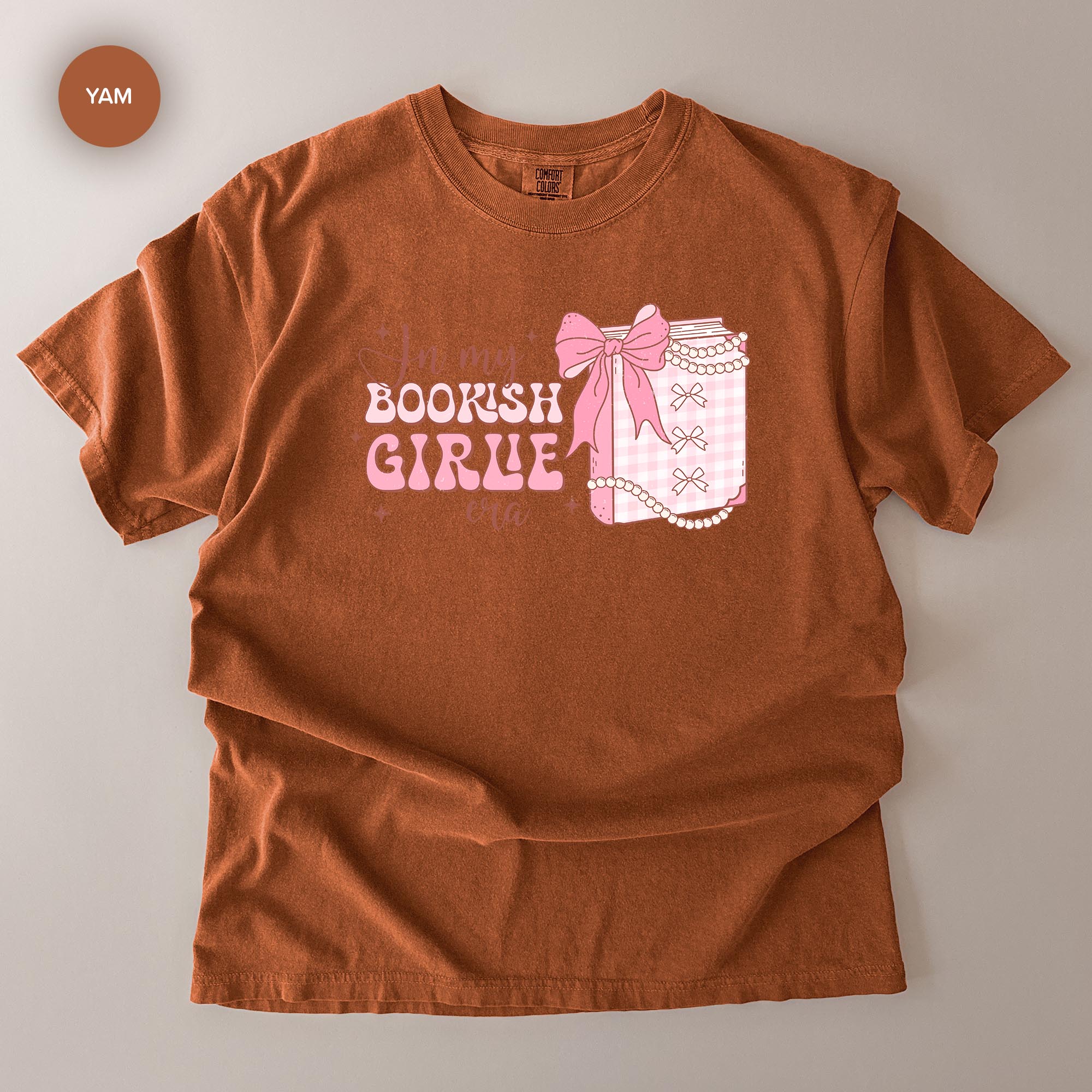 A brown t-shirt with a graphic design featuring a bow and a book, along with the text "In my BOOKISH GIRLE" printed on it.