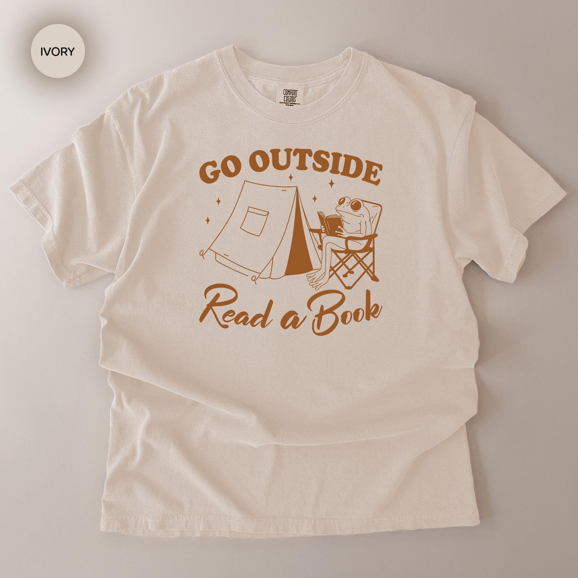 A white t-shirt with a graphic design that says "Go Outside Read a Book" and features a frog sitting in a chair next to a tent.