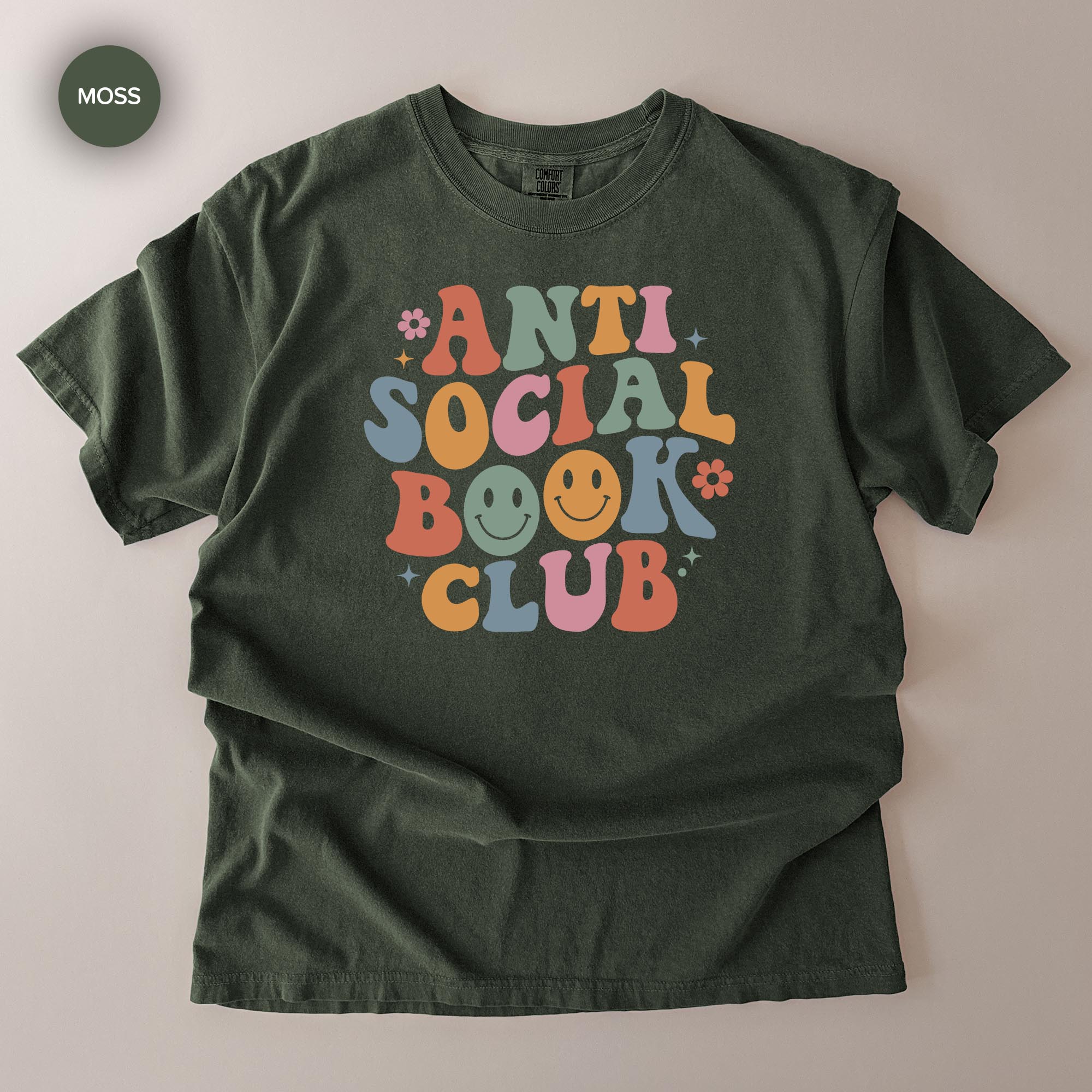 A green t-shirt with a colorful graphic design that says "ANTI SOCIAL BOOK CLUB" with smiley faces and flowers surrounding the text.