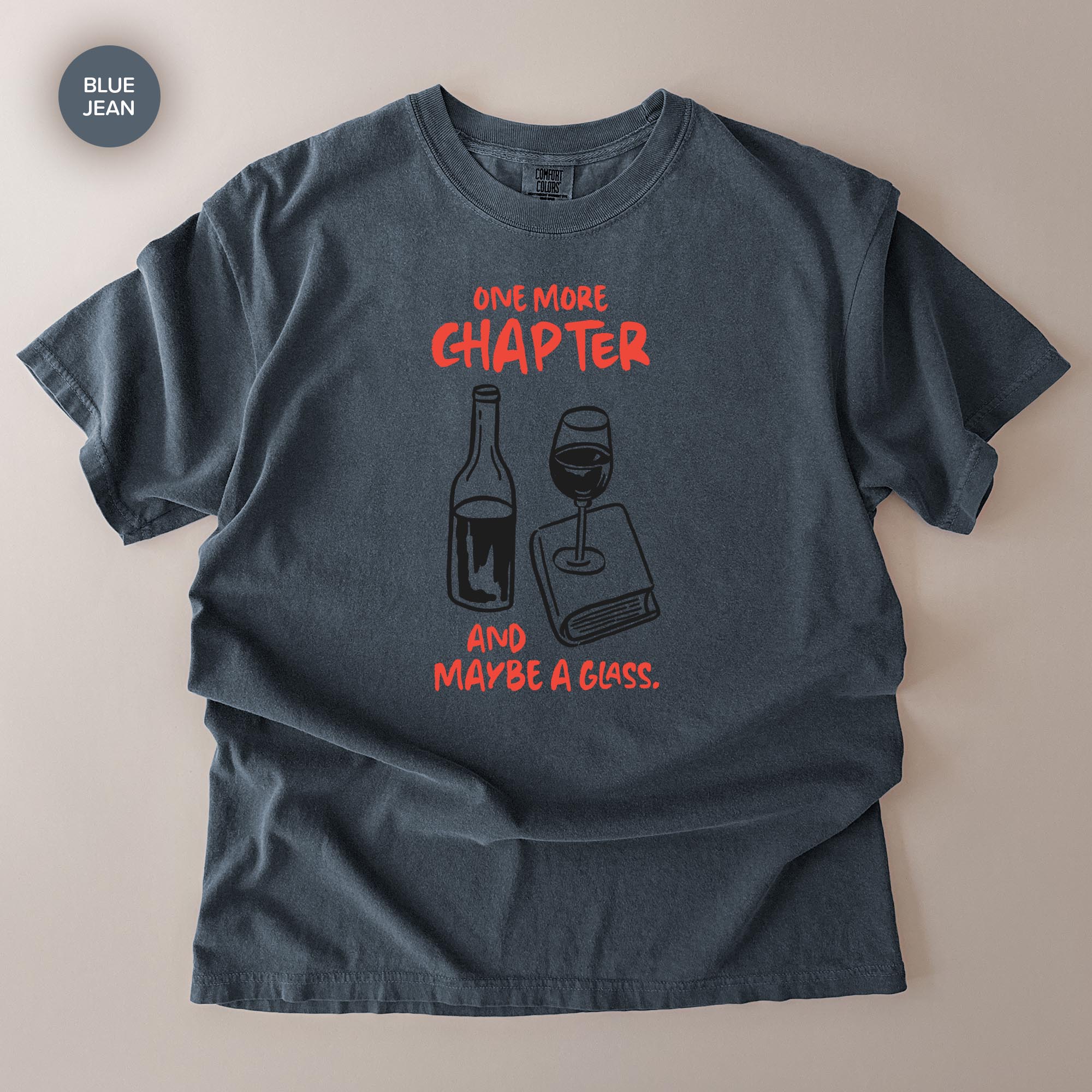 A gray t-shirt with a graphic design featuring a wine glass and a book, accompanied by the text "One more chapter and maybe a glass."