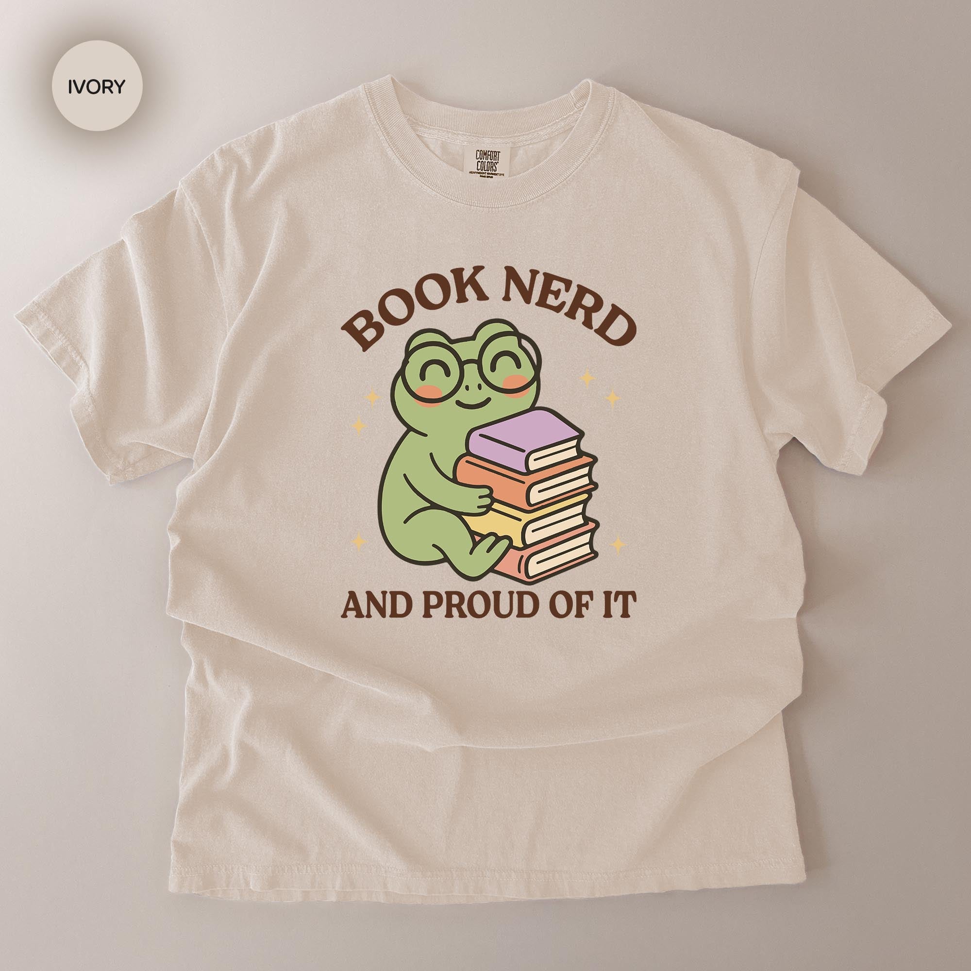 A white t-shirt with a graphic design featuring a frog wearing glasses and holding a stack of books, along with the text "BOOK NERD AND PROUD OF IT".