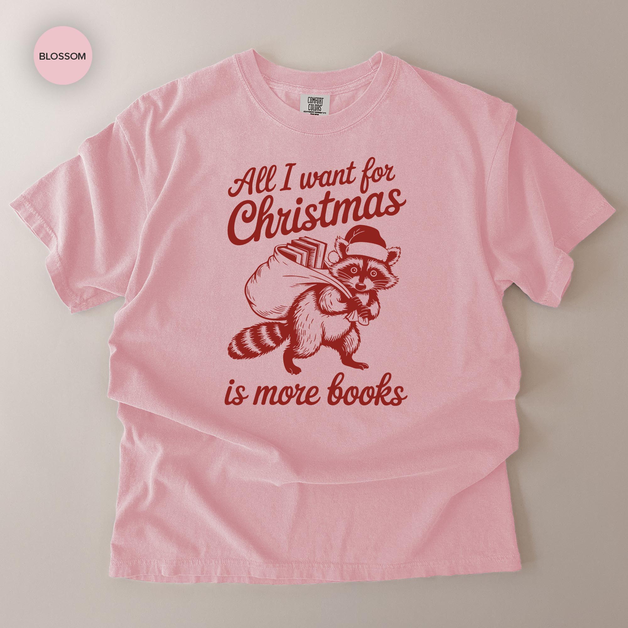 A pink t-shirt with a graphic design depicting a raccoon carrying a sack of books, accompanied by the text "All I want for Christmas is more books".
