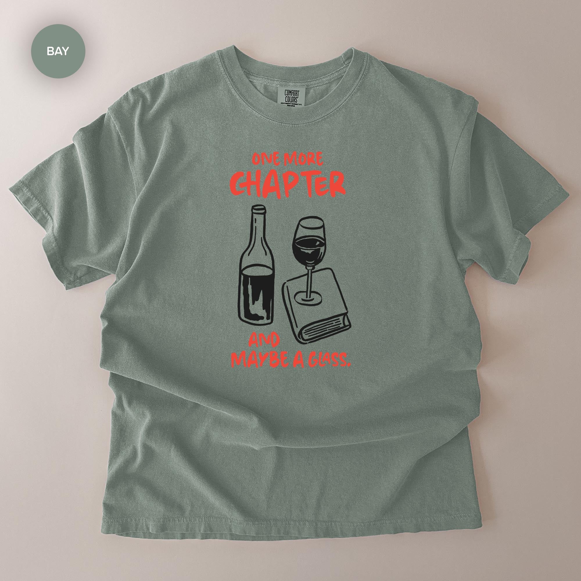 A gray t-shirt with a graphic design depicting a wine glass and a book, accompanied by the text "One More Chapter and maybe a glass of wine."