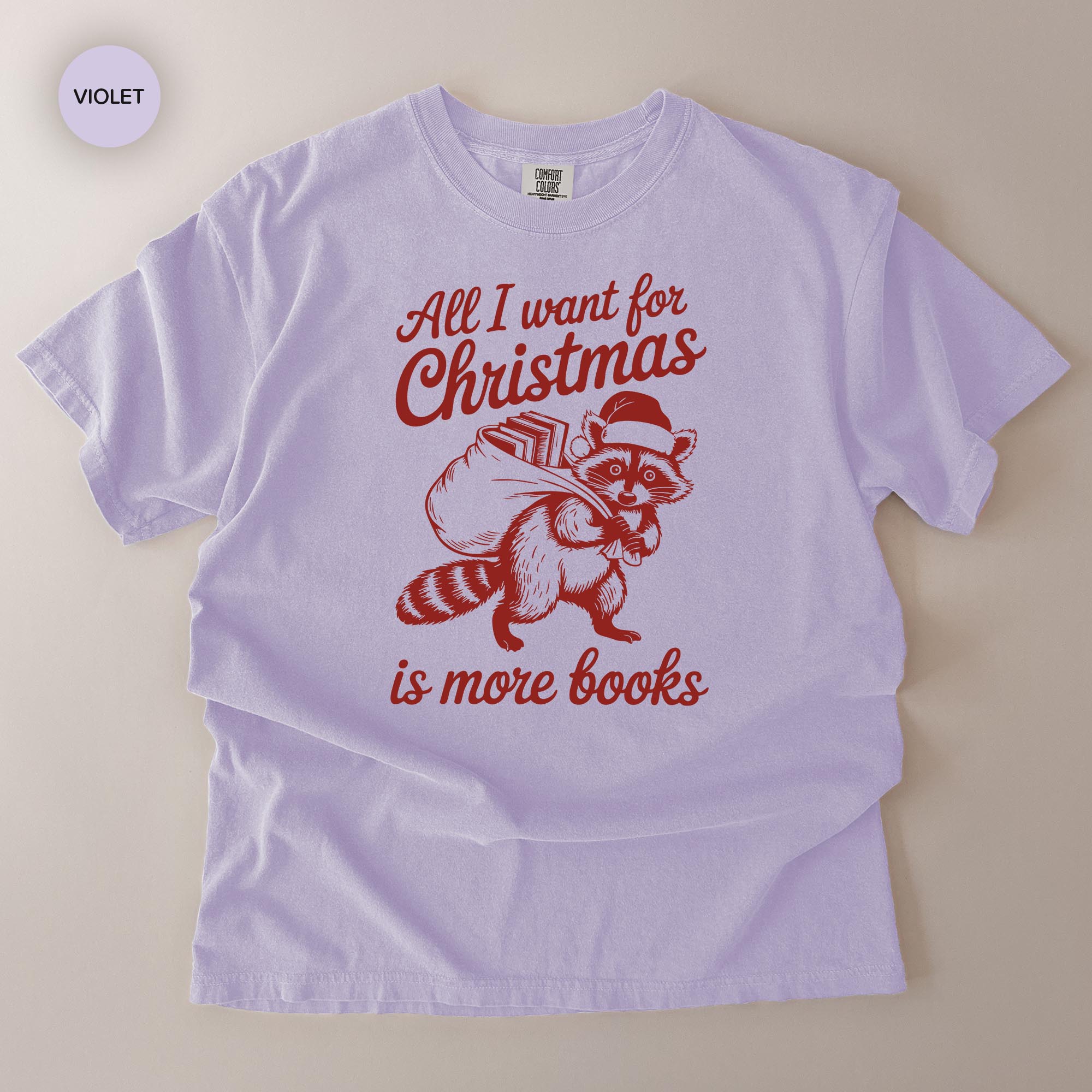 A white t-shirt with a red graphic design depicting a raccoon carrying a sack of books, along with the text "All I want for Christmas is more books".