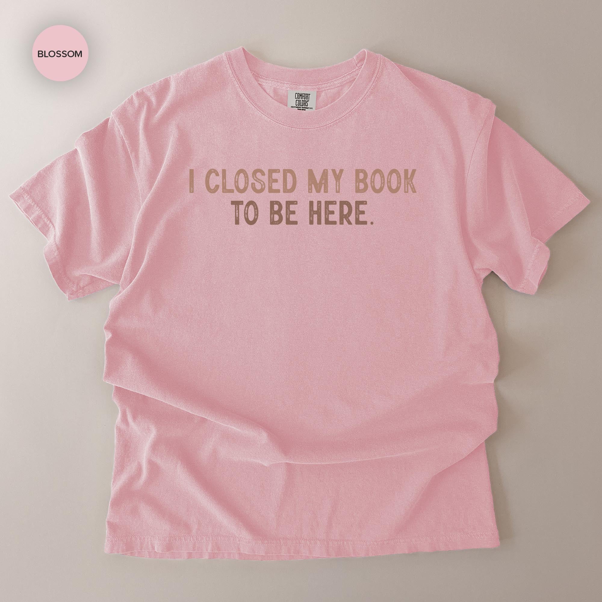 A pink t-shirt with the text "I CLOSED MY BOOK TO BE HERE" printed on it.