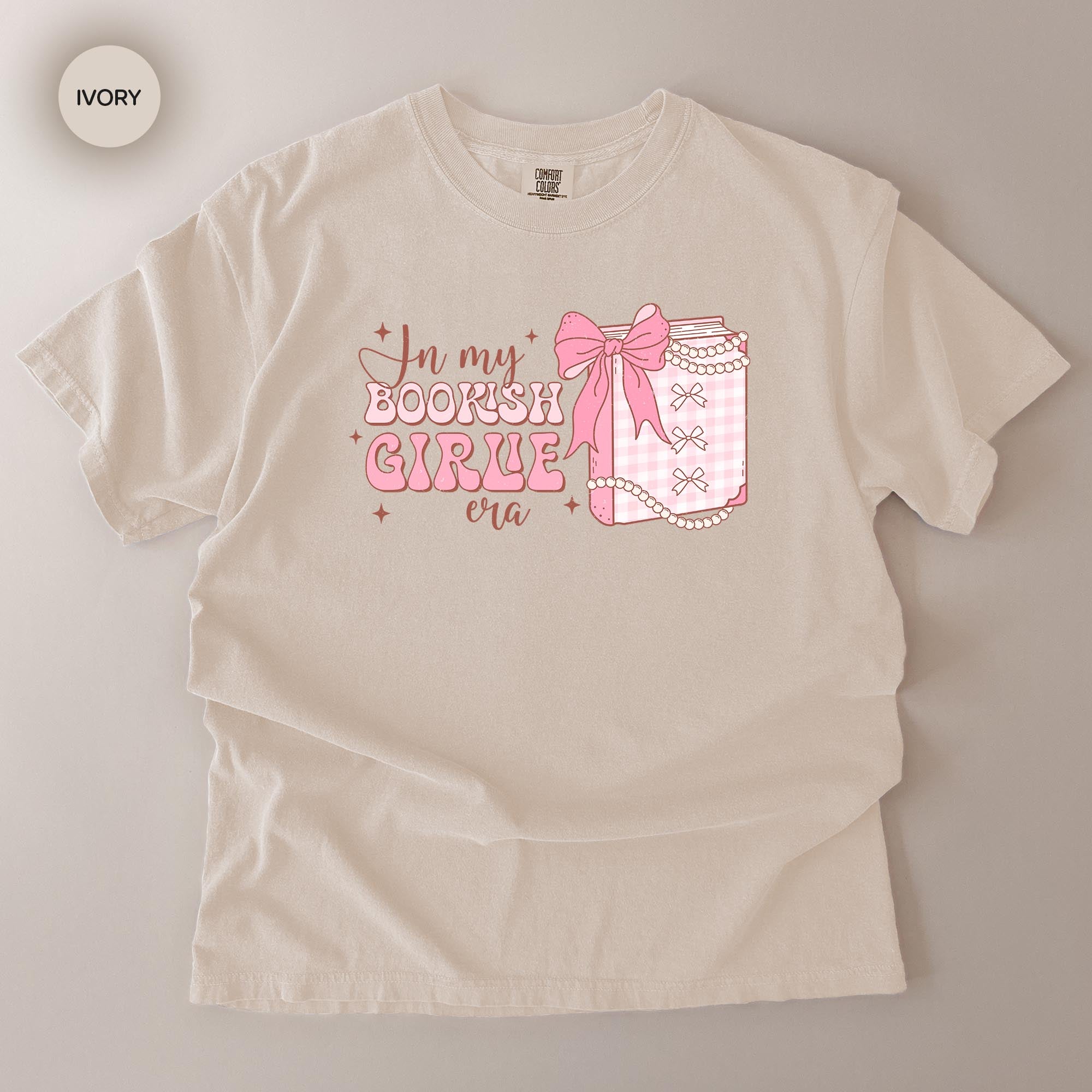 A white t-shirt with a graphic design featuring a bow and text that reads "In my BOOKSHOT GIRLE era".