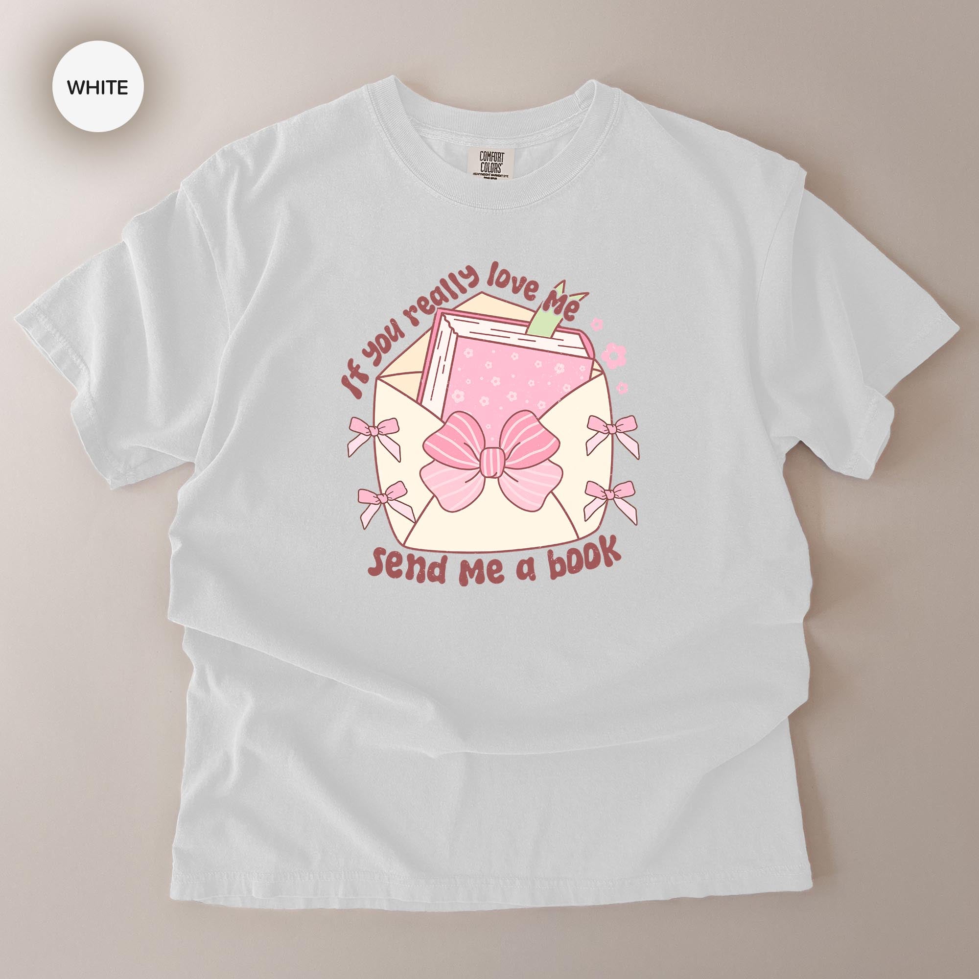 A white t-shirt with a graphic design featuring a book, bow, and text that reads "If you really love me, send me a book".