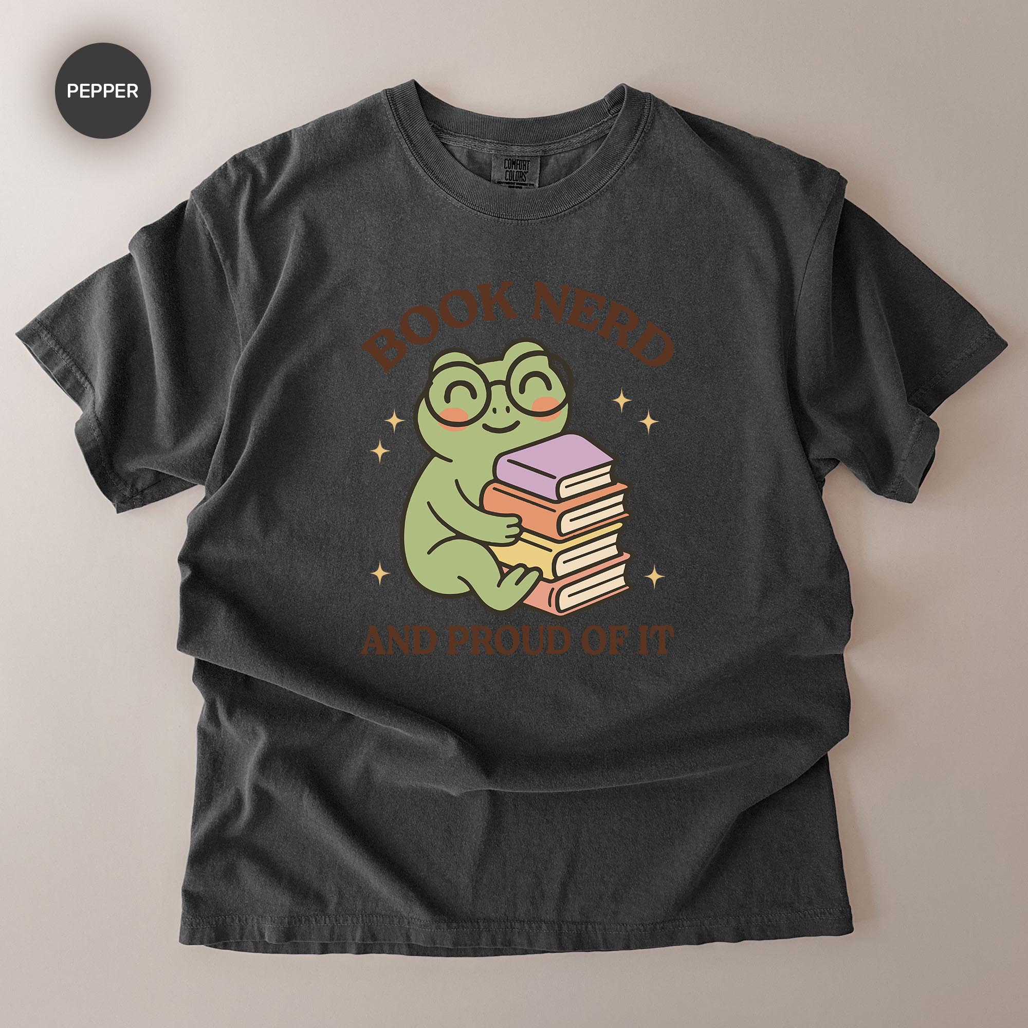 A black t-shirt with a graphic design featuring a smiling frog holding a stack of books, surrounded by stars. The text above the frog reads "BOOK NERD" and below the frog reads "AND PROUD OF IT".