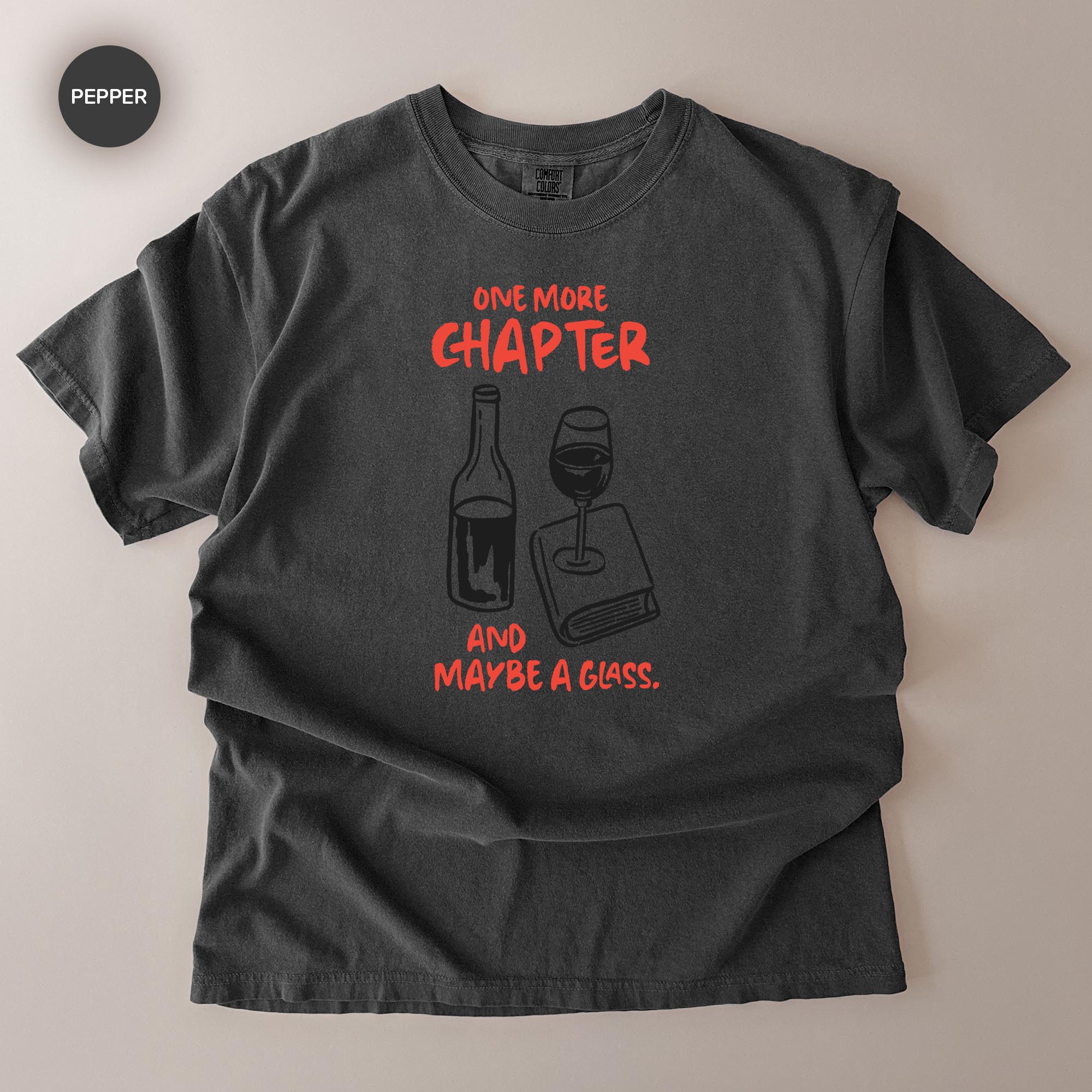 A black t-shirt with a graphic design featuring a wine glass and a book, accompanied by the text "ONE MORE CHAPTER AND MAYBE A GLASS."