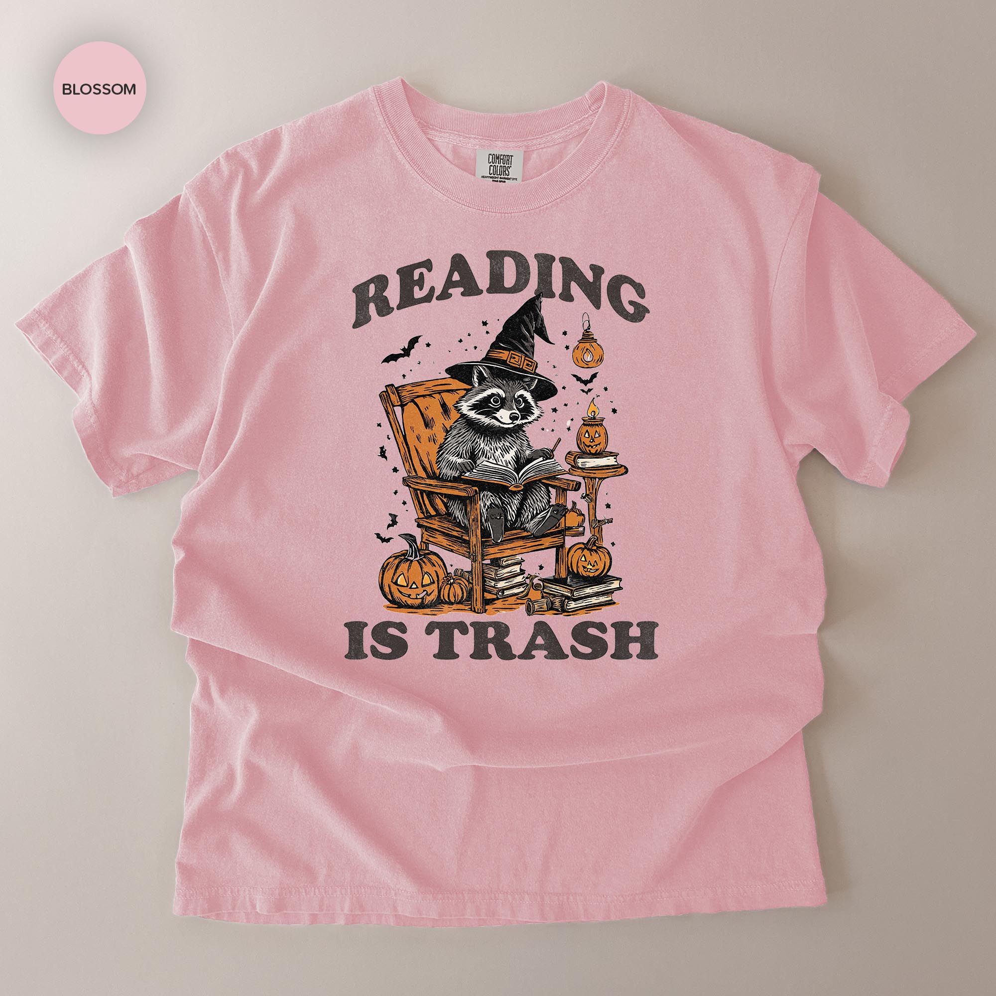 A pink t-shirt with a graphic design featuring a raccoon wearing a witch's hat and reading a book, surrounded by Halloween-themed elements such as pumpkins and bats.