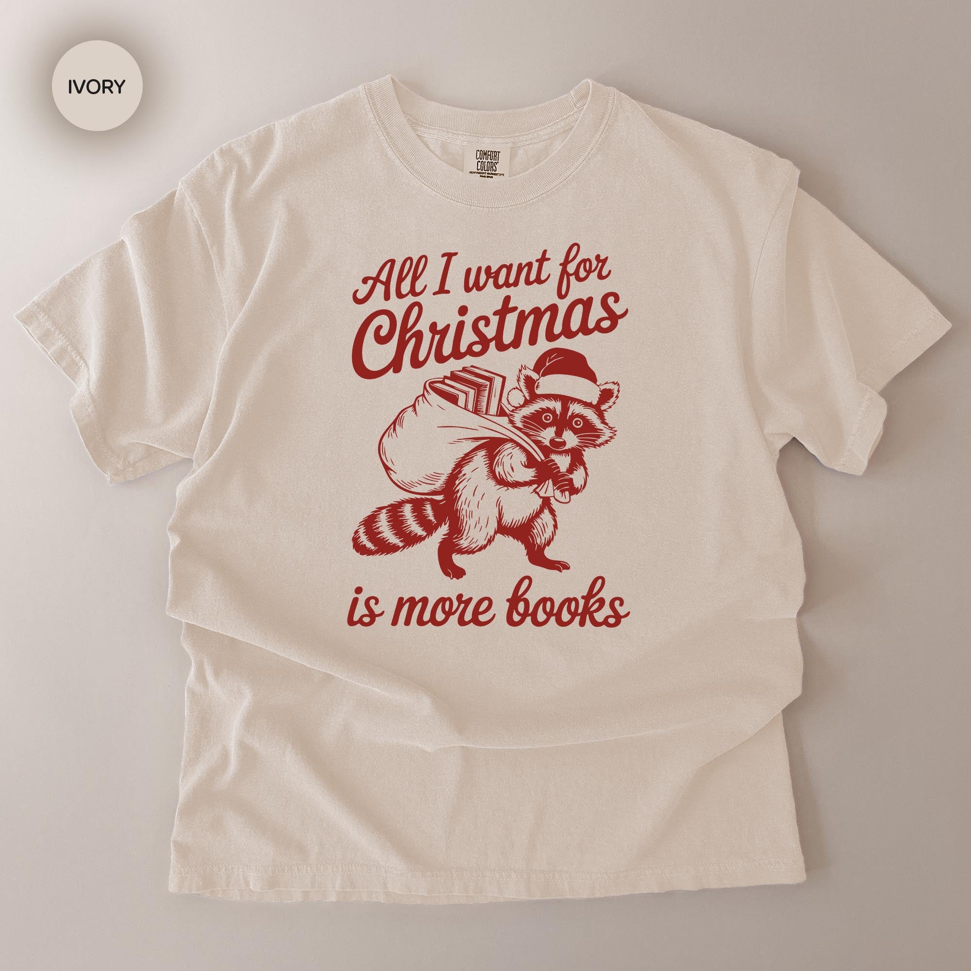 A white t-shirt with a red graphic design depicting a raccoon carrying a sack of books and the text "All I want for Christmas is more books".