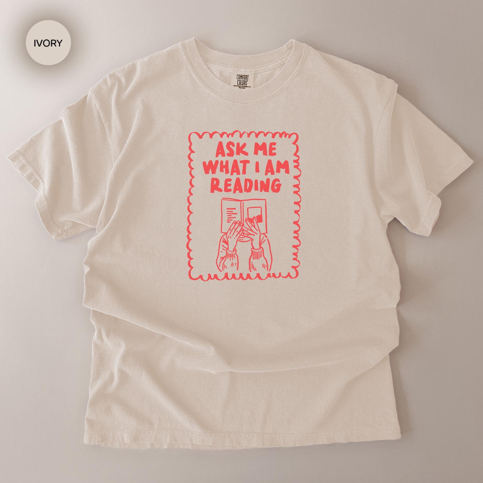 a white t-shirt with a red graphic design on it. The design includes a person reading a book and the text "Ask me what I am reading".