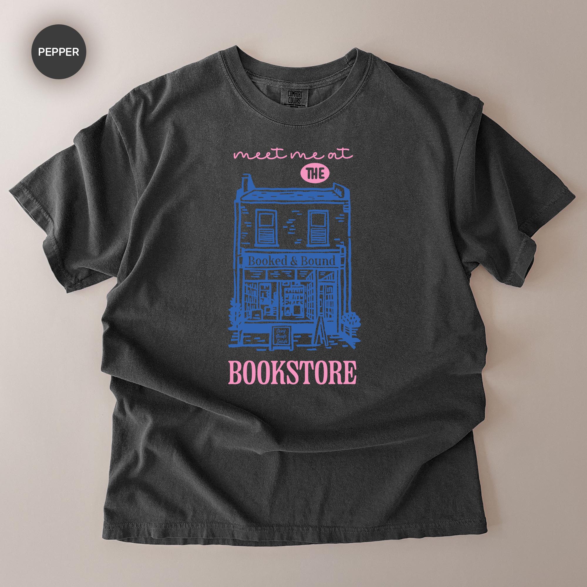 A gray t-shirt with a graphic design depicting a building and the text "Meet me at THE BOOKSTORE" printed on it.