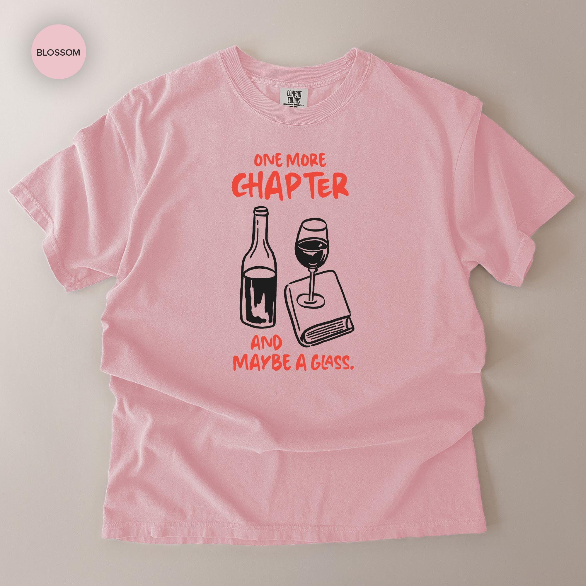 A pink t-shirt with a graphic design depicting a wine glass and a book, accompanied by the text "One more chapter and maybe a glass."