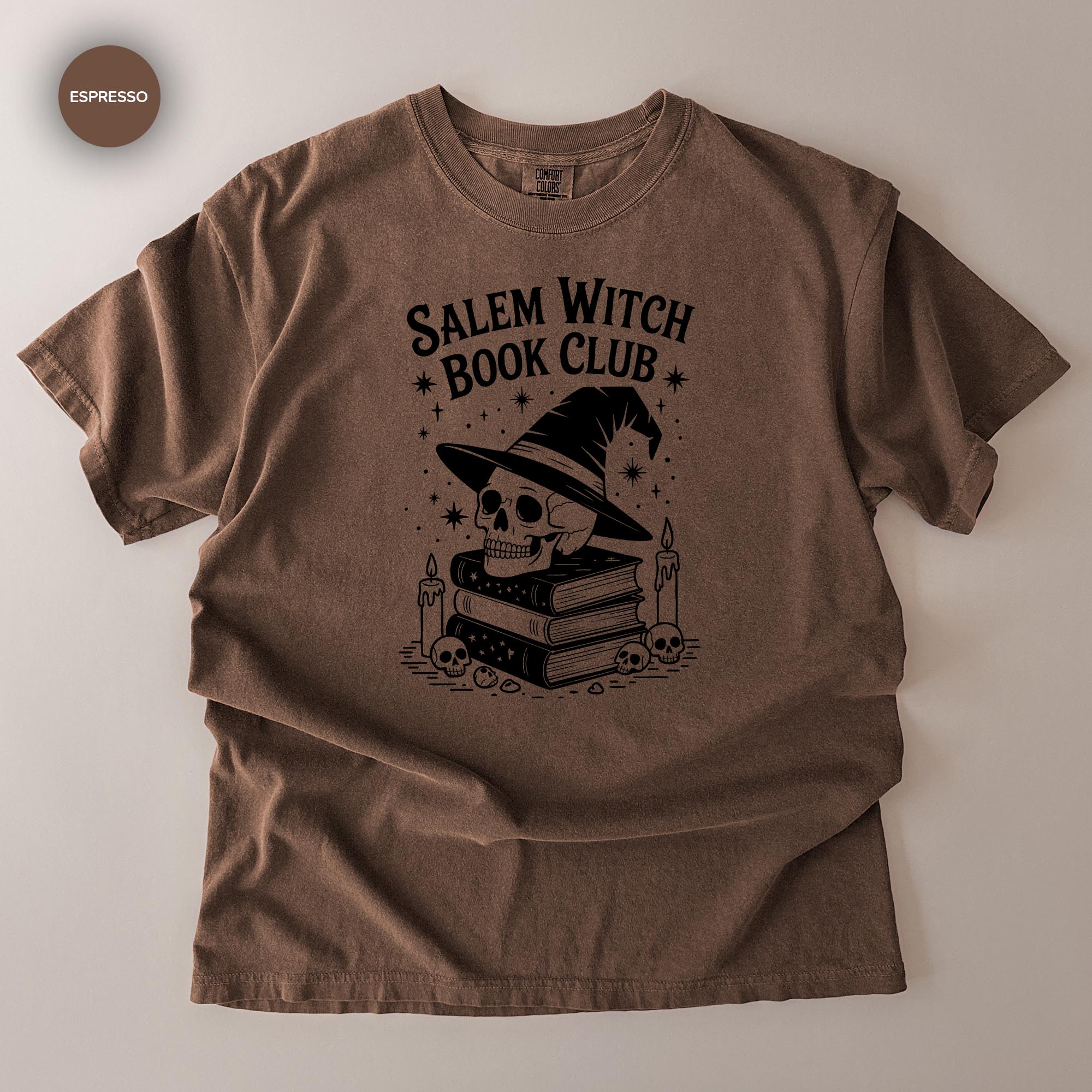 A brown t-shirt with a graphic design featuring a skull wearing a witch's hat, surrounded by books and candles, with the text "Salem Witch Book Club" printed on it.