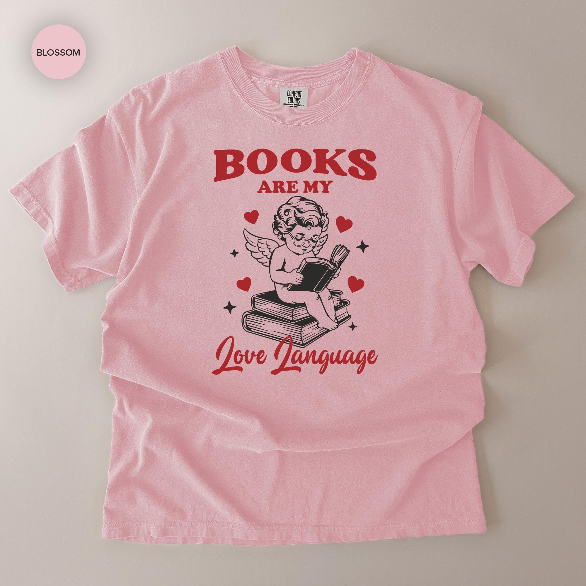 A pink t-shirt with a graphic design featuring a cherub holding a book and surrounded by hearts, along with the text "BOOKS ARE MY LOVE LANGUAGE".