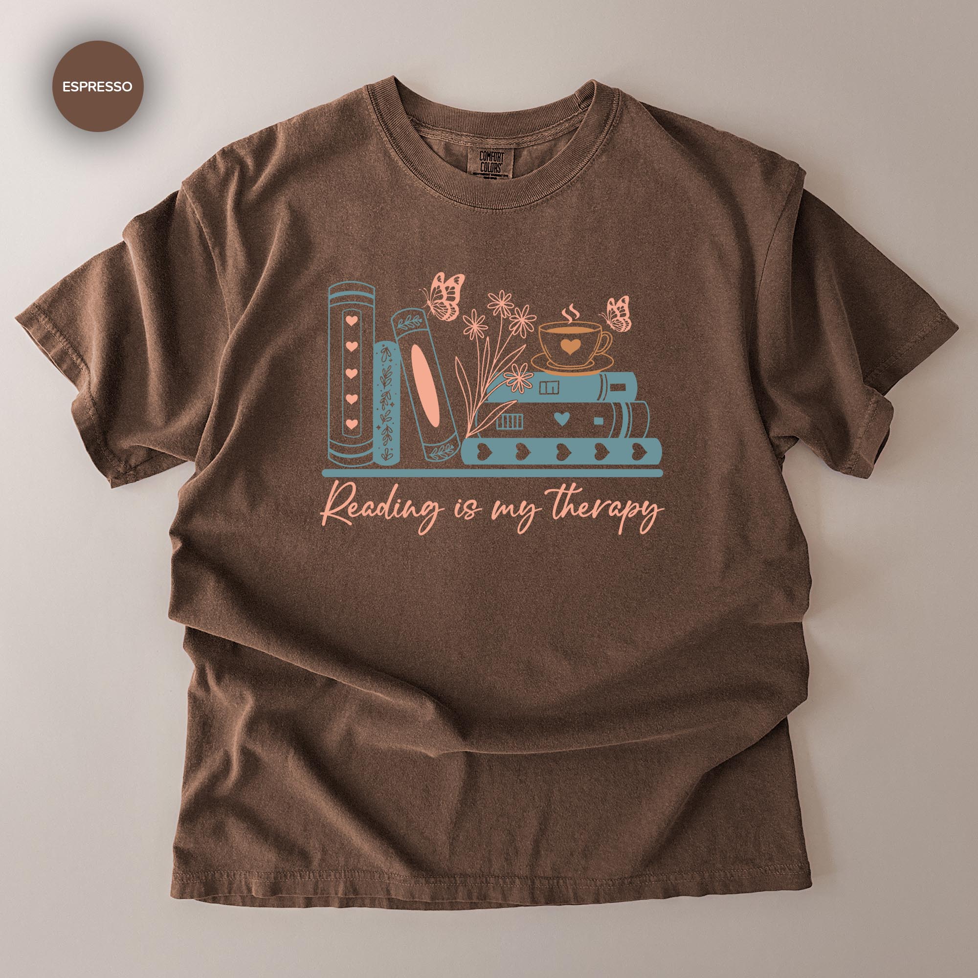 A brown t-shirt with a graphic design featuring books, flowers, and butterflies, along with the text "Reading is my therapy".
