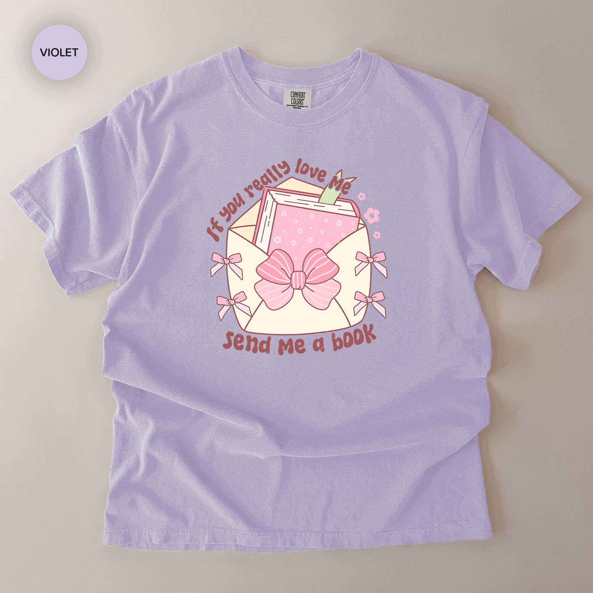 A purple t-shirt with a graphic design featuring a bow, a book, and the text "If you really love me, send me a book".