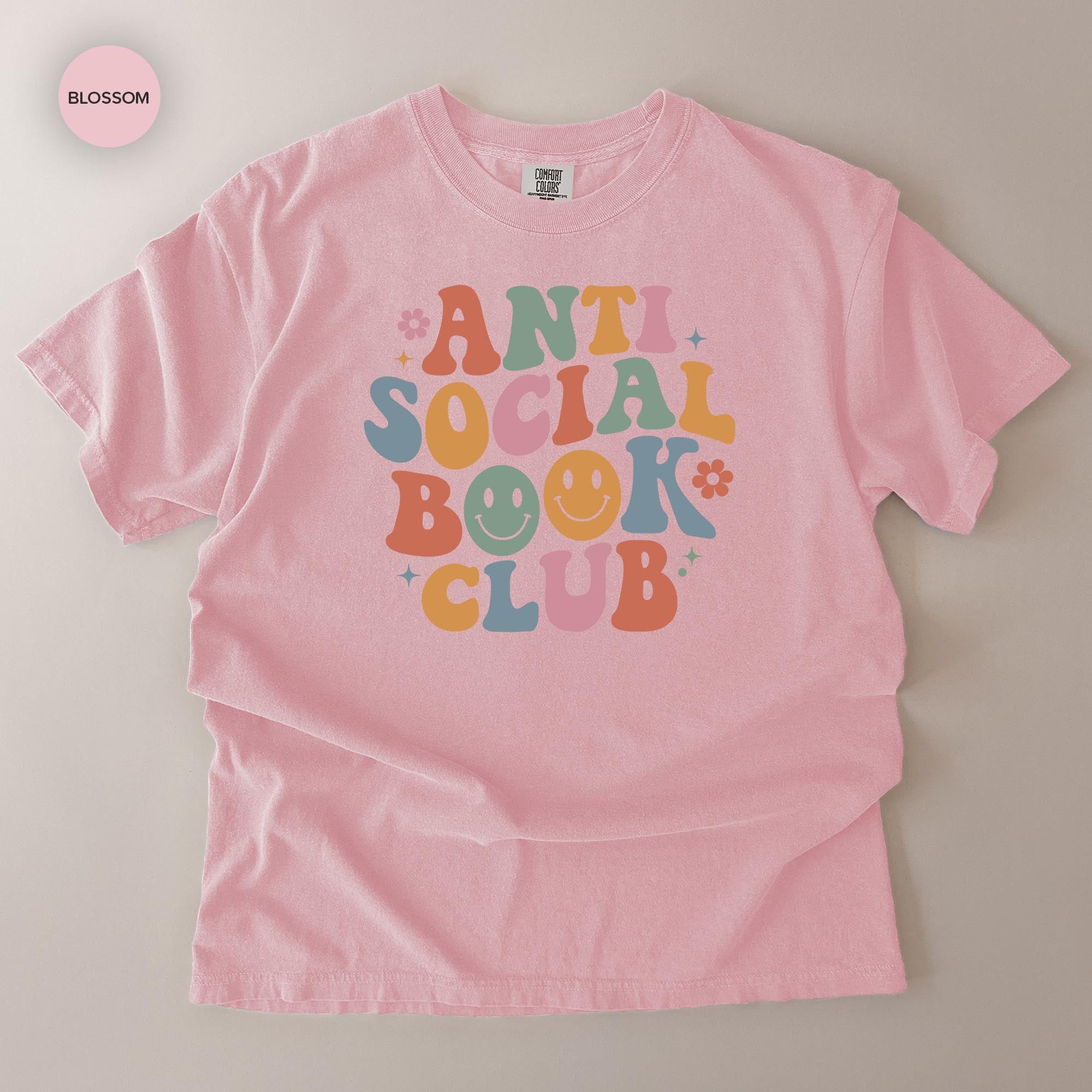 A pink t-shirt with a colorful graphic design that says "ANTI SOCIAL BOOK CLUB" in a playful, retro style.