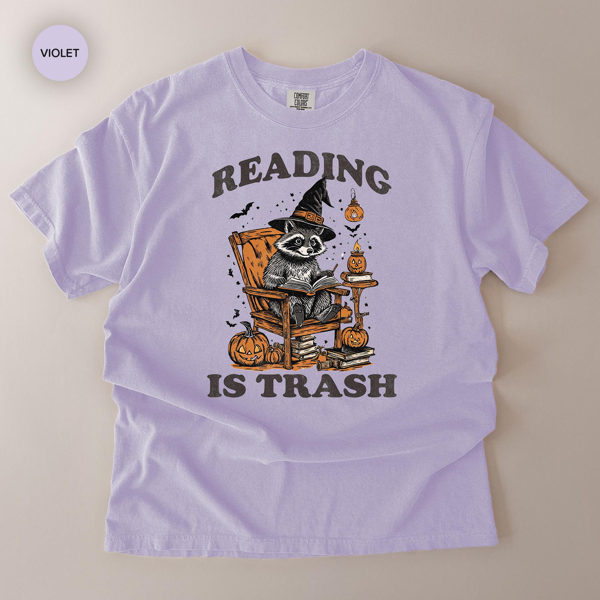 A purple t-shirt with a graphic design depicting a raccoon wearing a witch's hat and reading a book, surrounded by Halloween-themed elements such as pumpkins and bats.