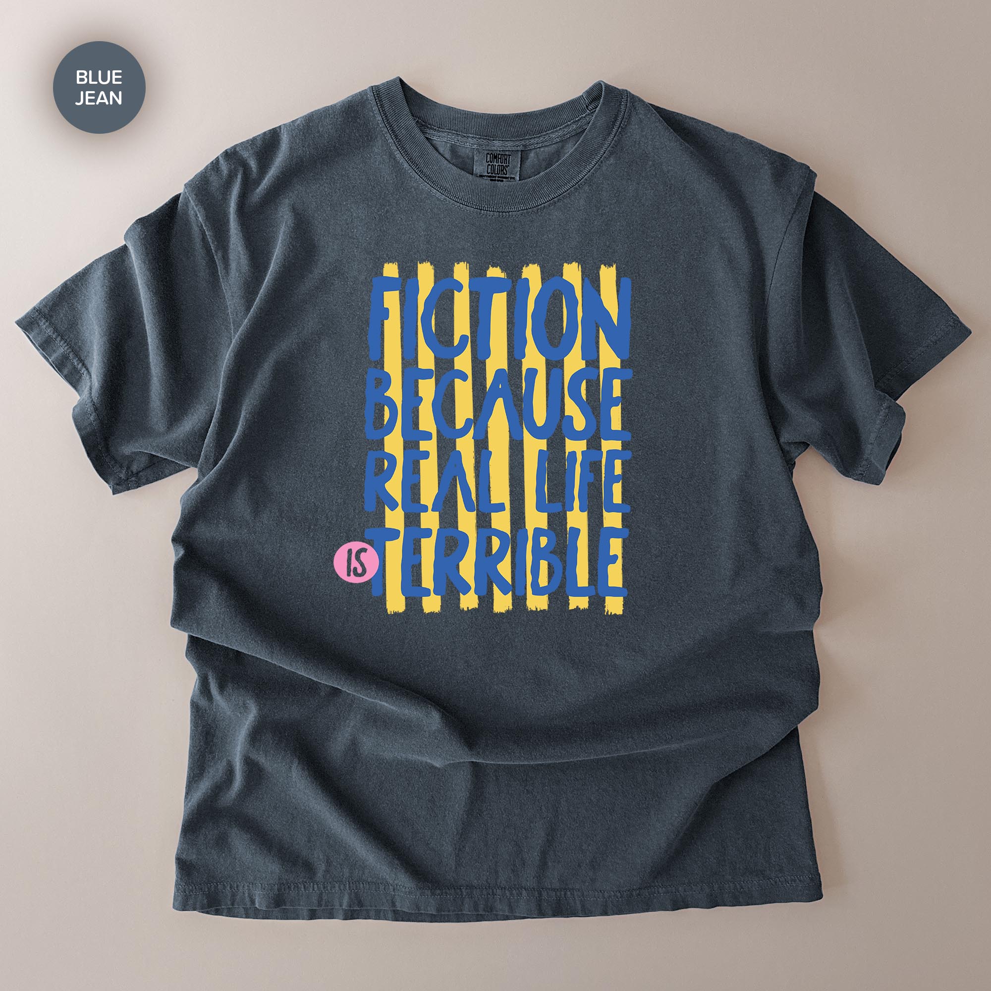 A gray t-shirt with a graphic design featuring the text "FICTION BECAUSE REAL LIFE IS TERRIBLE" in blue and yellow colors.