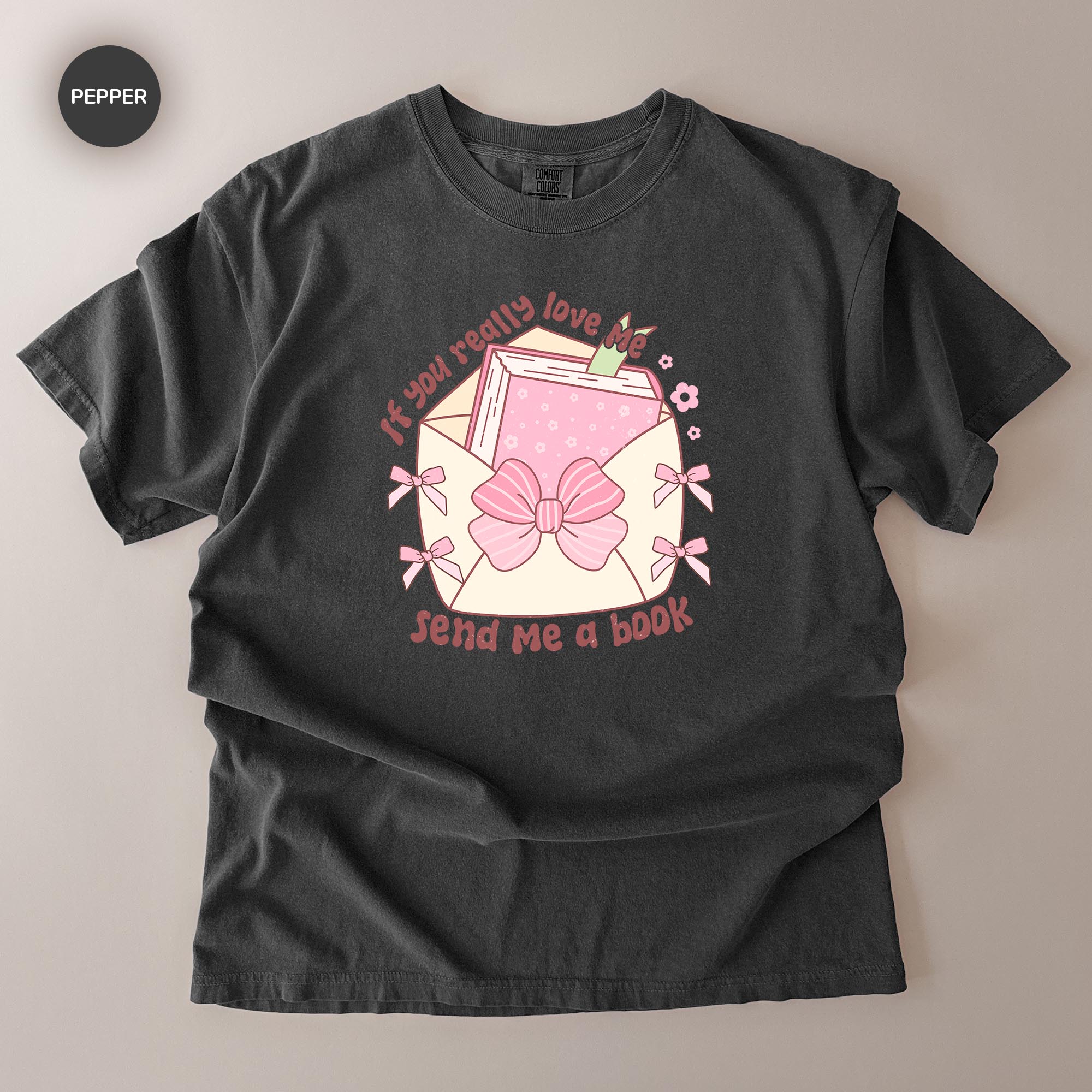 A black t-shirt with a graphic design featuring a pink bow, a book, and the text "If you really love me, send me a book".