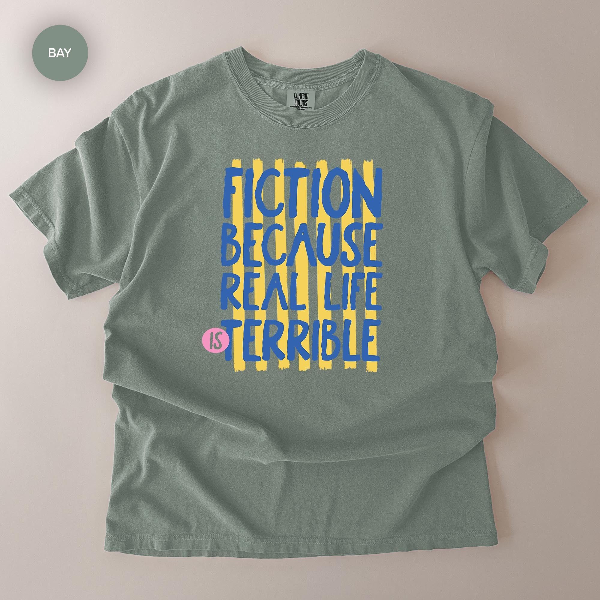 A gray t-shirt with a graphic design featuring the text "FICTION BECAUSE REAL LIFE IS TERRIBLE" in blue and yellow colors.