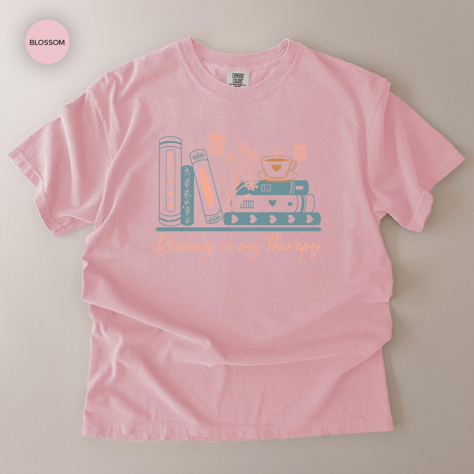 A pink t-shirt with a graphic design featuring a stack of books and a cup of tea, along with the text "Reading is my therapy".