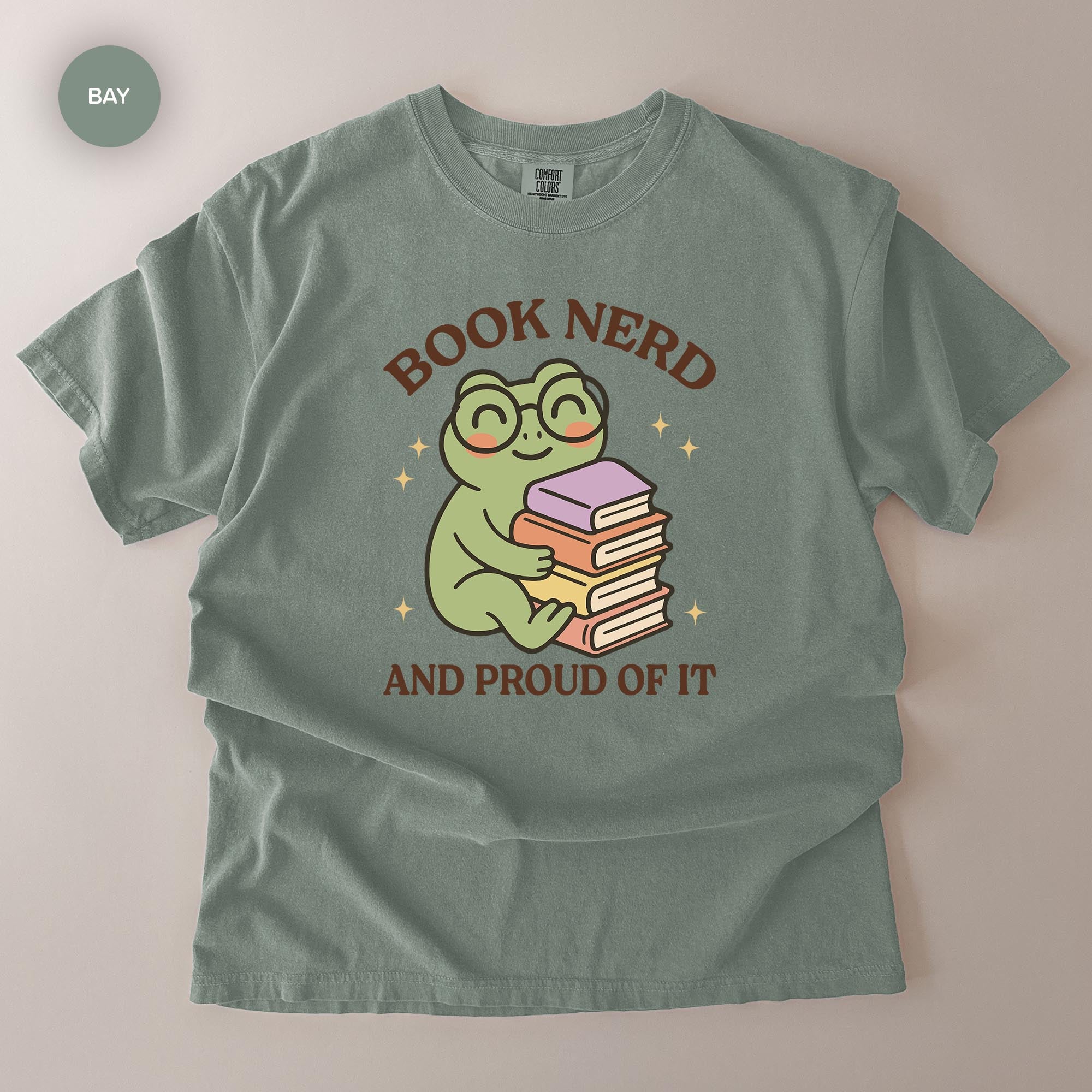 A gray t-shirt with a graphic design featuring a frog wearing glasses and holding a stack of books, along with the text "BOOK NERD AND PROUD OF IT".