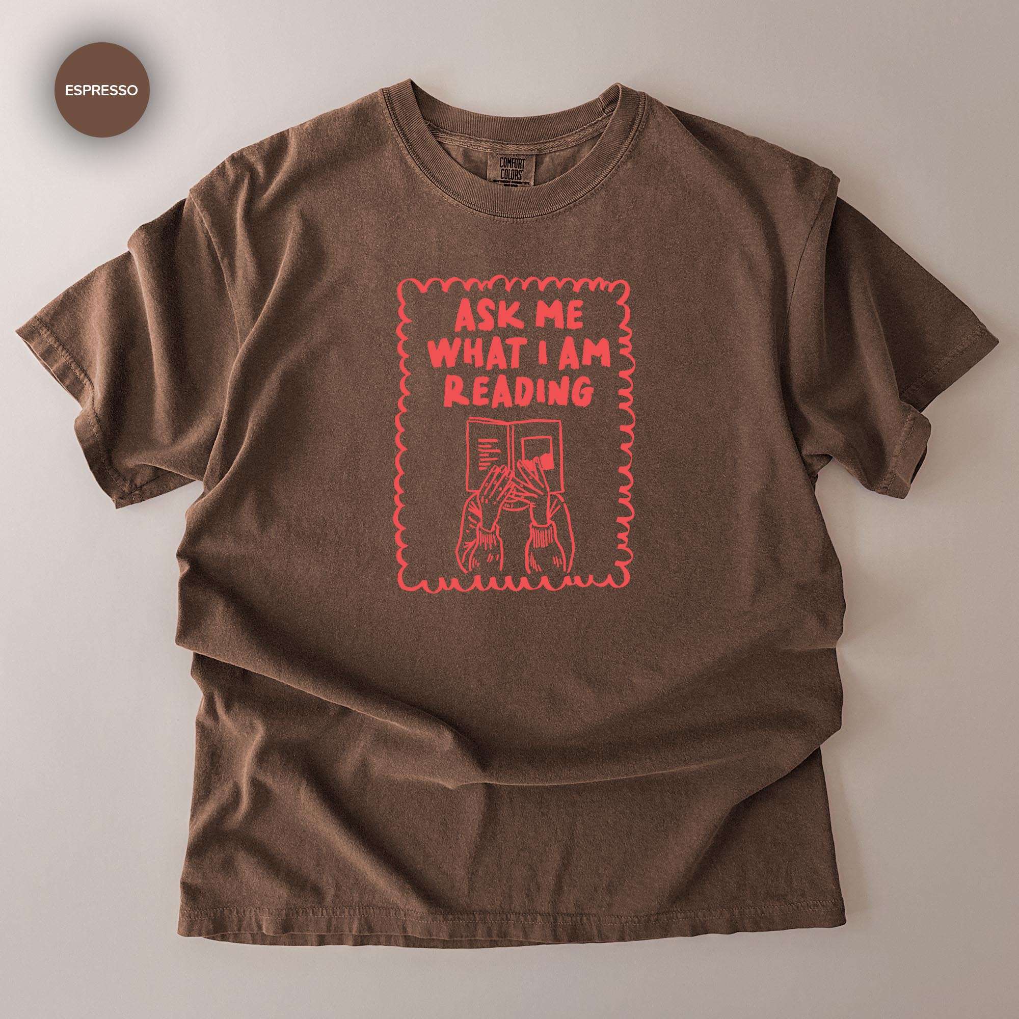 a brown t-shirt with a graphic design on it. The design includes a person holding a book and the text "ask me what I am reading".