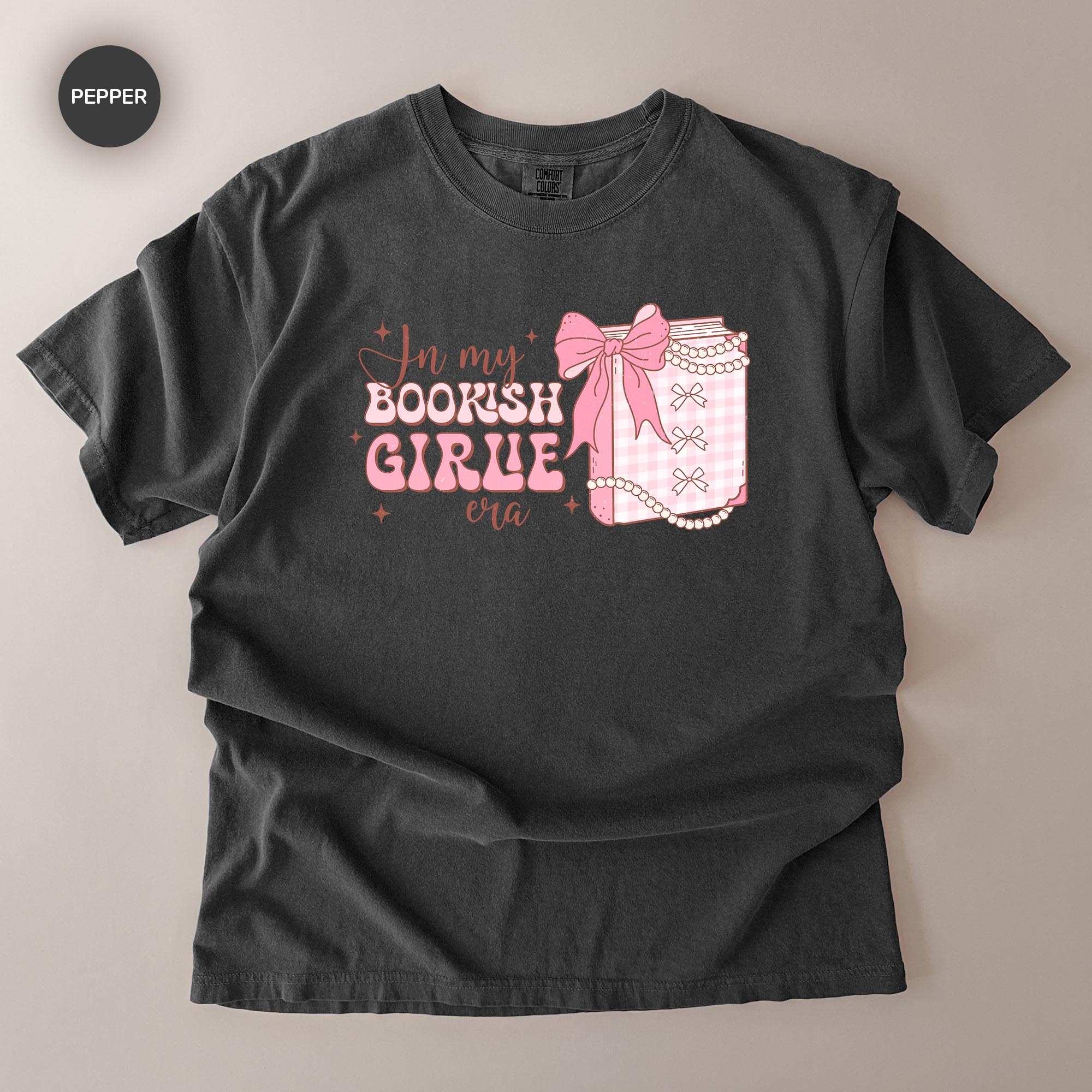 A black t-shirt with a graphic design featuring a bow, pearls, and text that reads "In my BOOKISH GIRLE era".