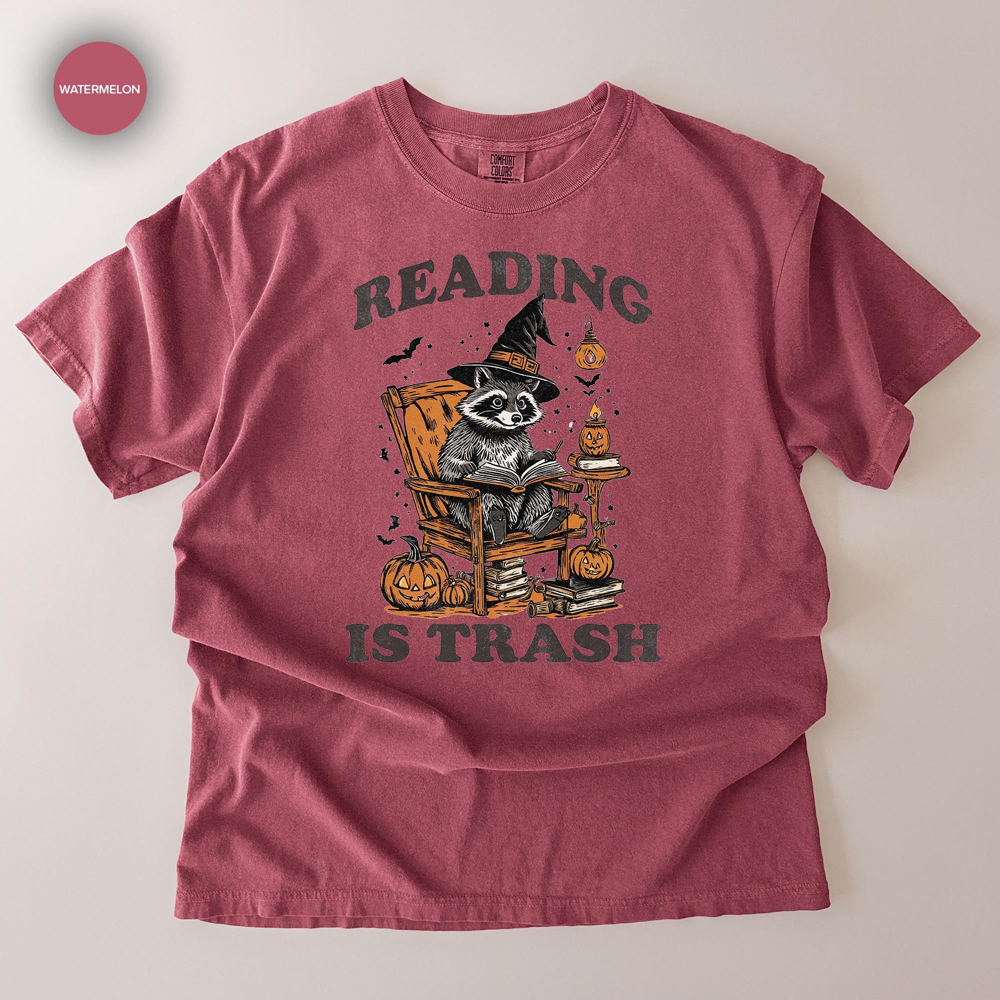 A red t-shirt with a graphic design depicting a raccoon wearing a witch's hat and reading a book, surrounded by Halloween-themed elements such as pumpkins and bats.