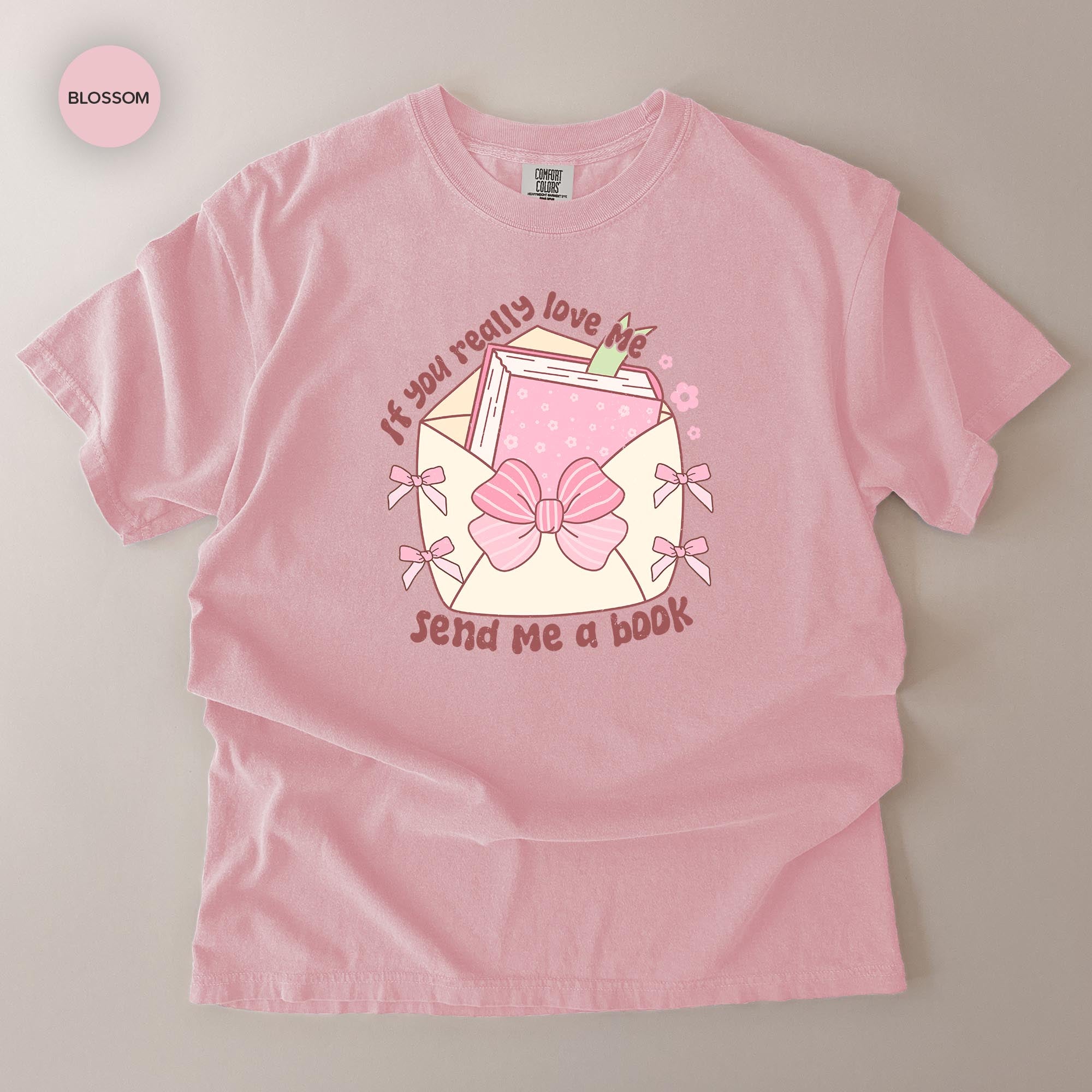 A pink t-shirt with a graphic design featuring a book, bows, and text that reads "If you really love me, send me a book".