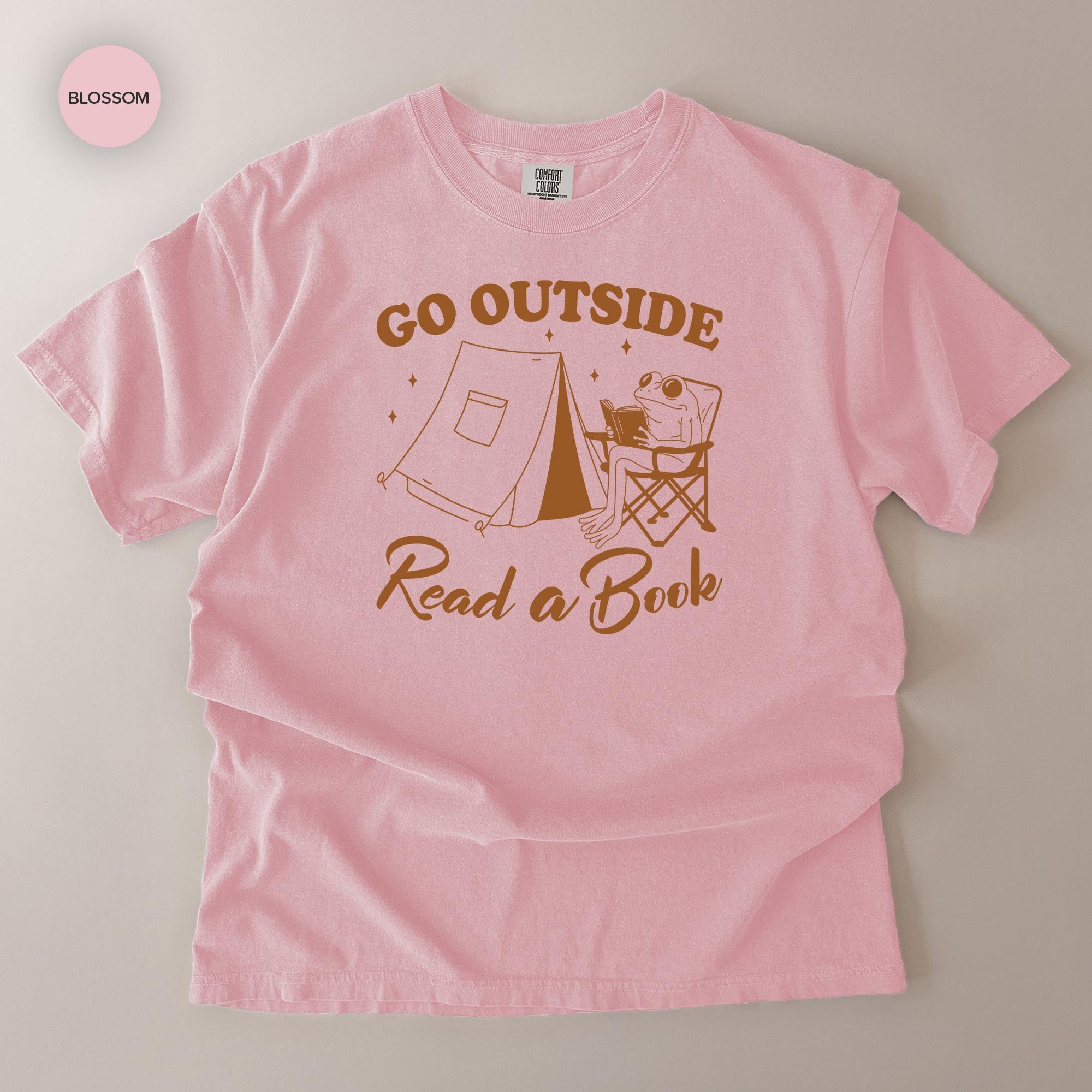 A pink t-shirt with a graphic design that says "Go Outside Read a Book" and features a frog sitting in a chair.