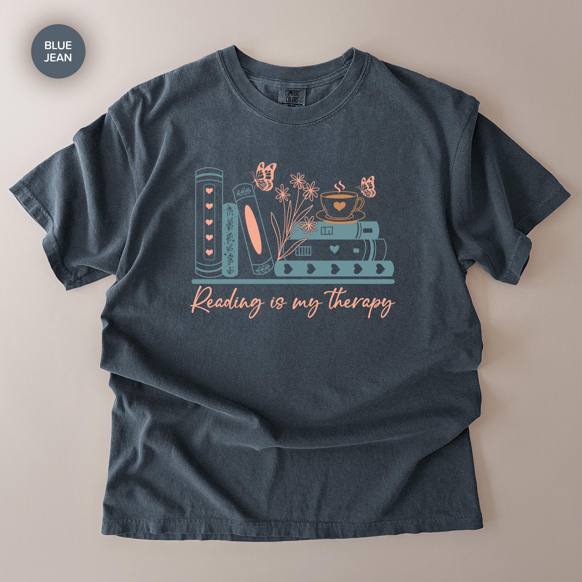 A gray t-shirt with a graphic design featuring a stack of books, flowers, butterflies, and the text "Reading is my therapy" printed on it.