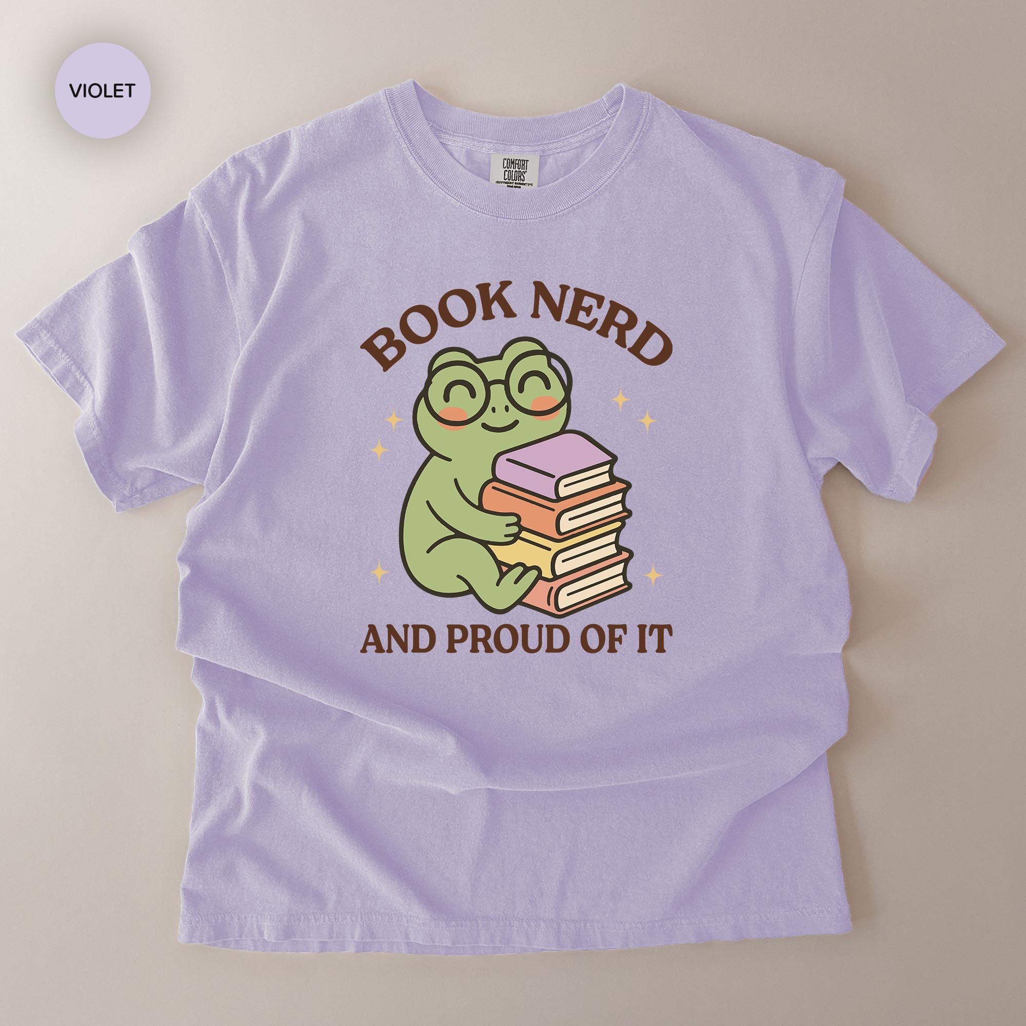 A purple t-shirt with a cartoon frog wearing glasses and holding a stack of books, along with the text "BOOK NERD AND PROUD OF IT".