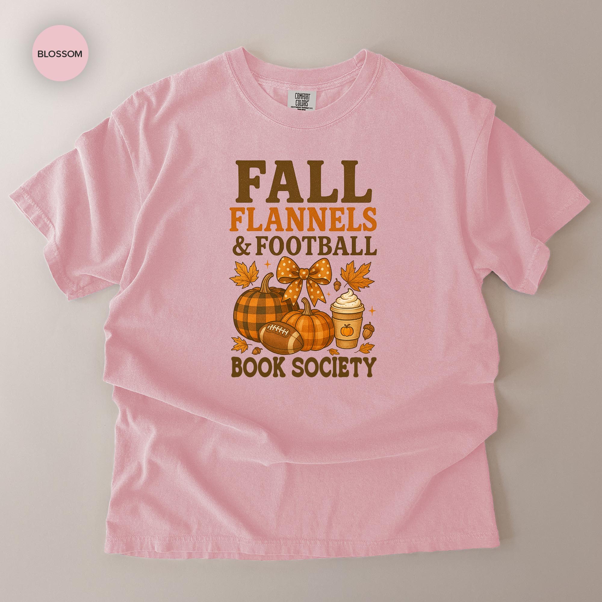 A pink t-shirt with a graphic design featuring fall-themed elements such as pumpkins, a football, and a cup of coffee, along with the text "FALL FLANNELS & FOOTBALL BOOK SOCIETY".