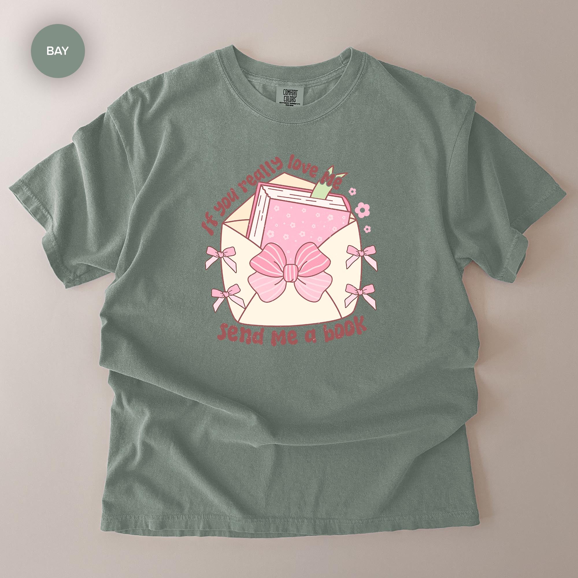 A gray t-shirt with a graphic design featuring a pink bow, a book, and the text "If you really love me, send me a book".