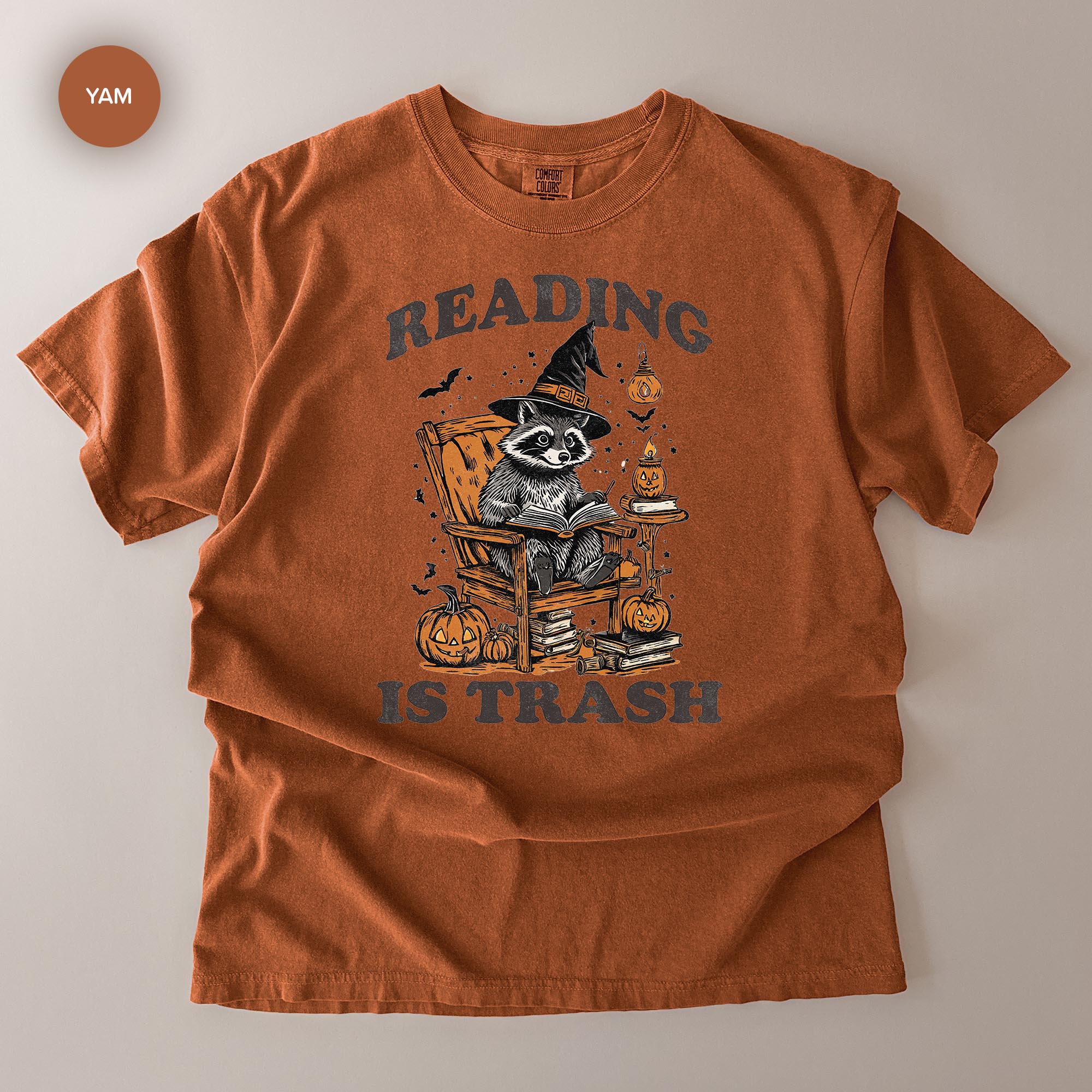 The image is a t-shirt with a graphic design featuring a raccoon wearing a witch's hat and holding a book, surrounded by Halloween-themed elements such as pumpkins and bats.