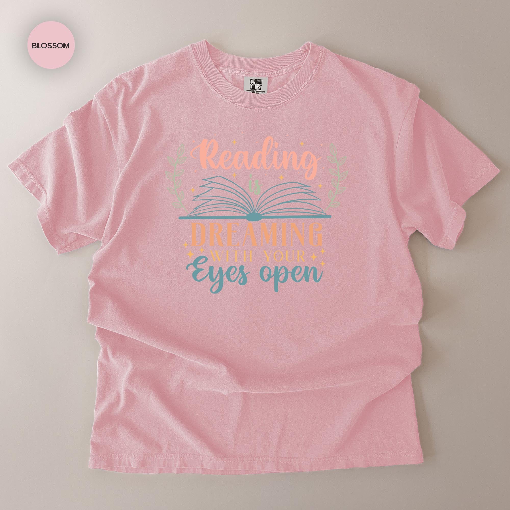 A pink t-shirt with a printed design that says "Reading Dreams With Eyes Open" and features an open book with a flower design.