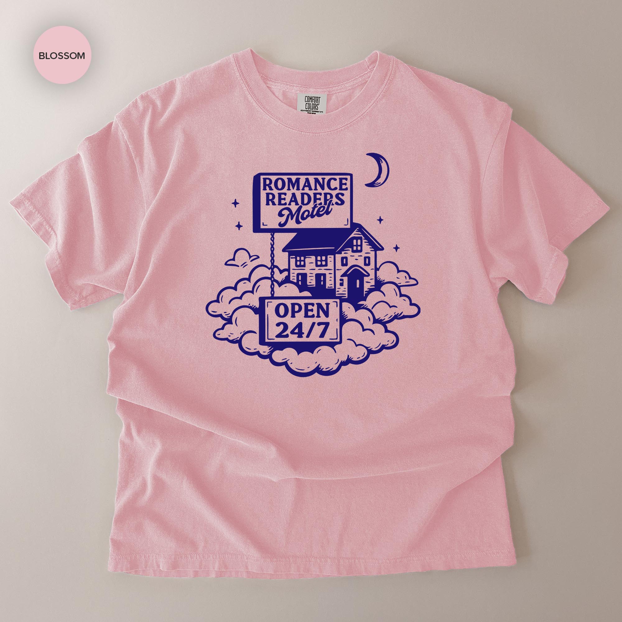 A pink t-shirt with a graphic design featuring a house, clouds, and a crescent moon, along with the text "ROMANCE READERS MOILET" and "OPEN 24/7".