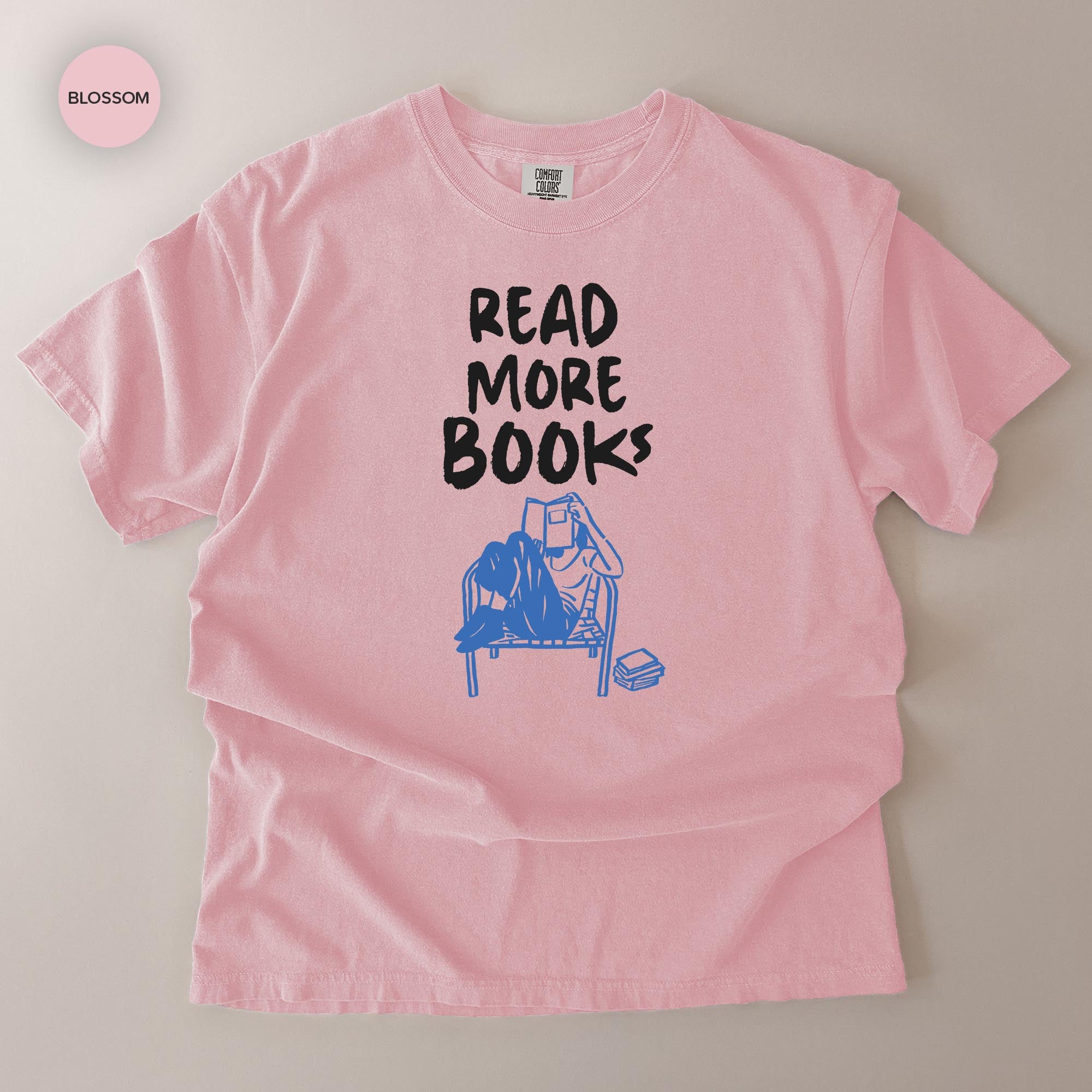 A pink t-shirt with the text "READ MORE BOOKS" printed on it, along with an illustration of a person reading a book.