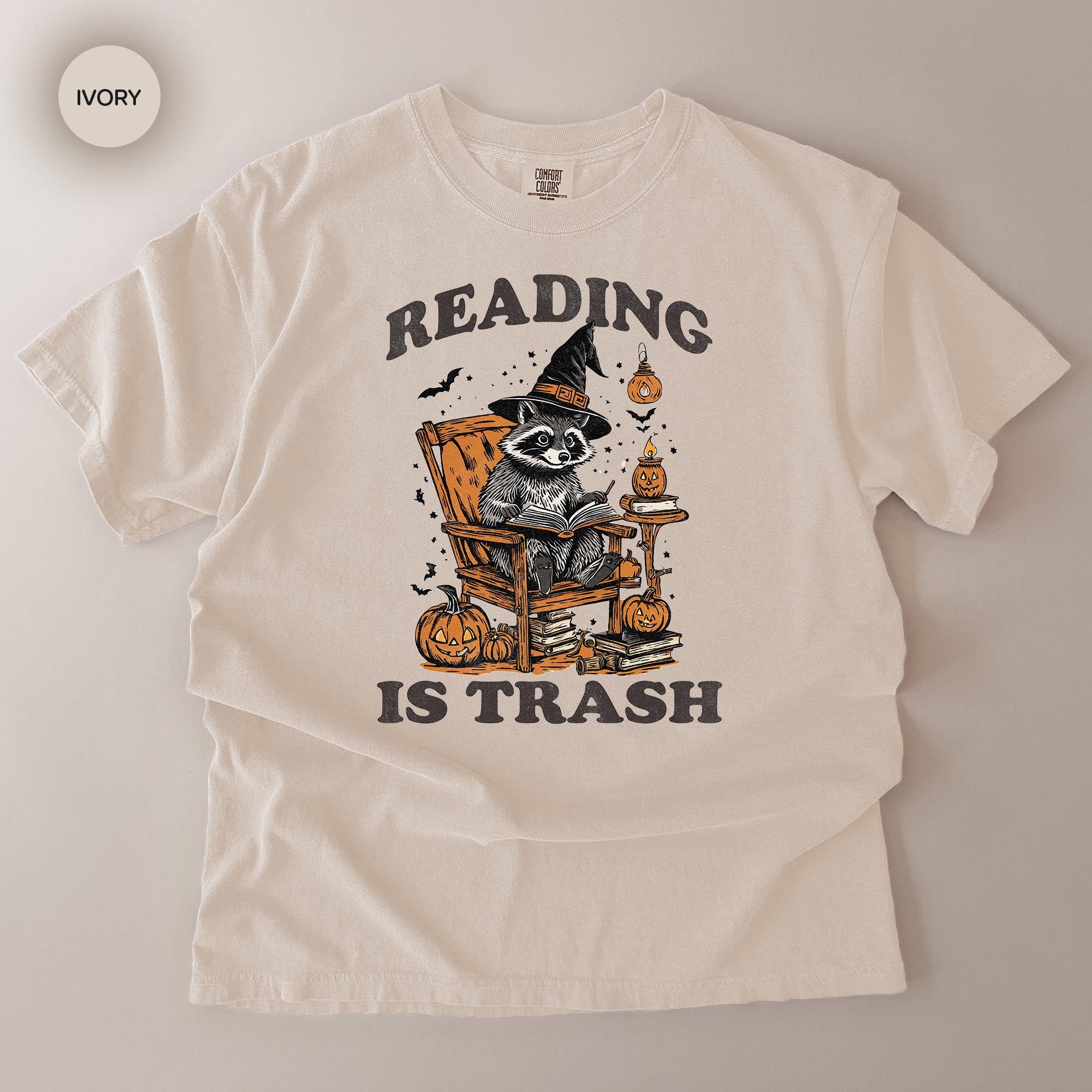 A white t-shirt with a graphic design featuring a raccoon wearing a witch's hat and holding a book, surrounded by Halloween-themed elements such as pumpkins and bats.