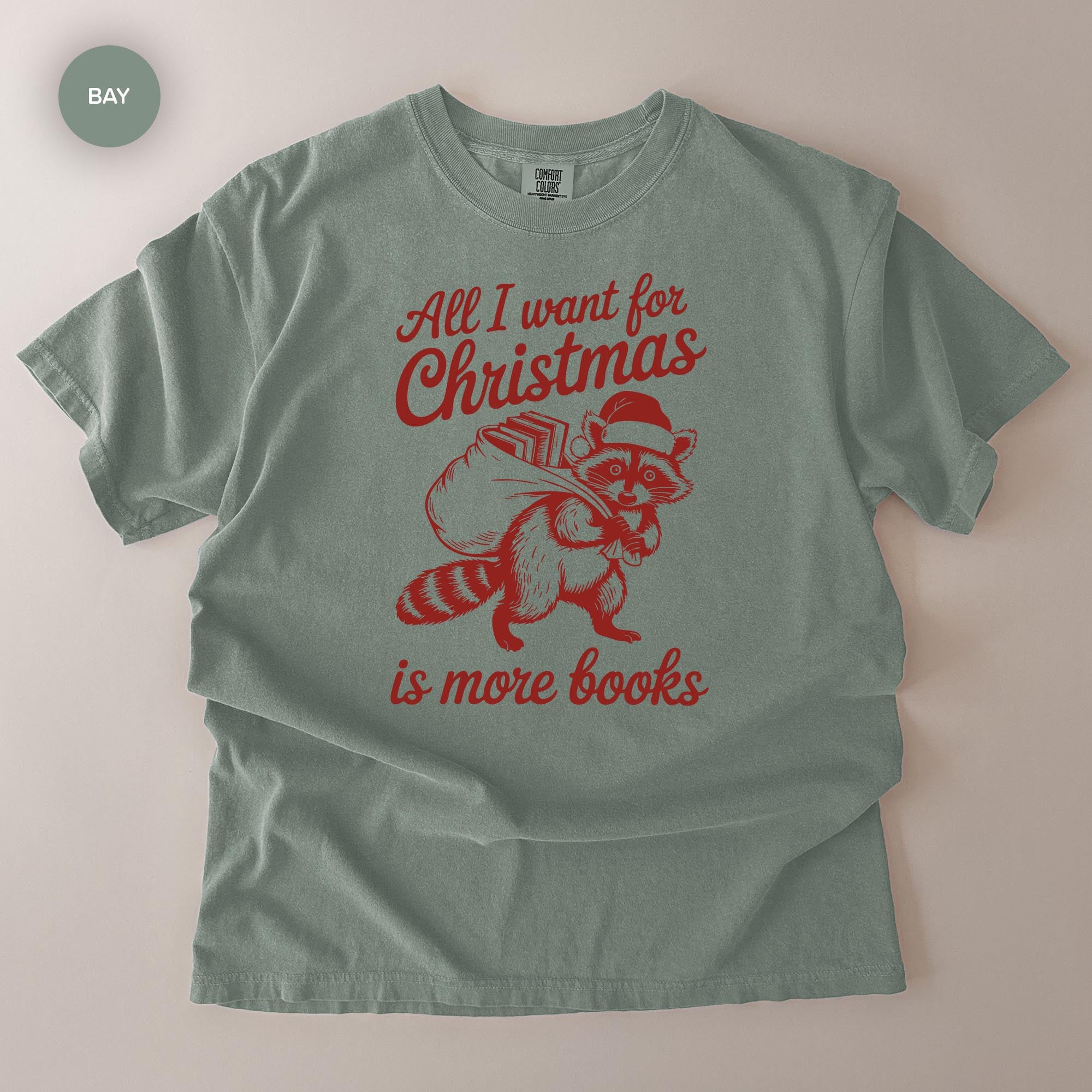 A gray t-shirt with a red graphic design depicting a raccoon carrying a sack of books and the text "All I want for Christmas is more books" printed on it.
