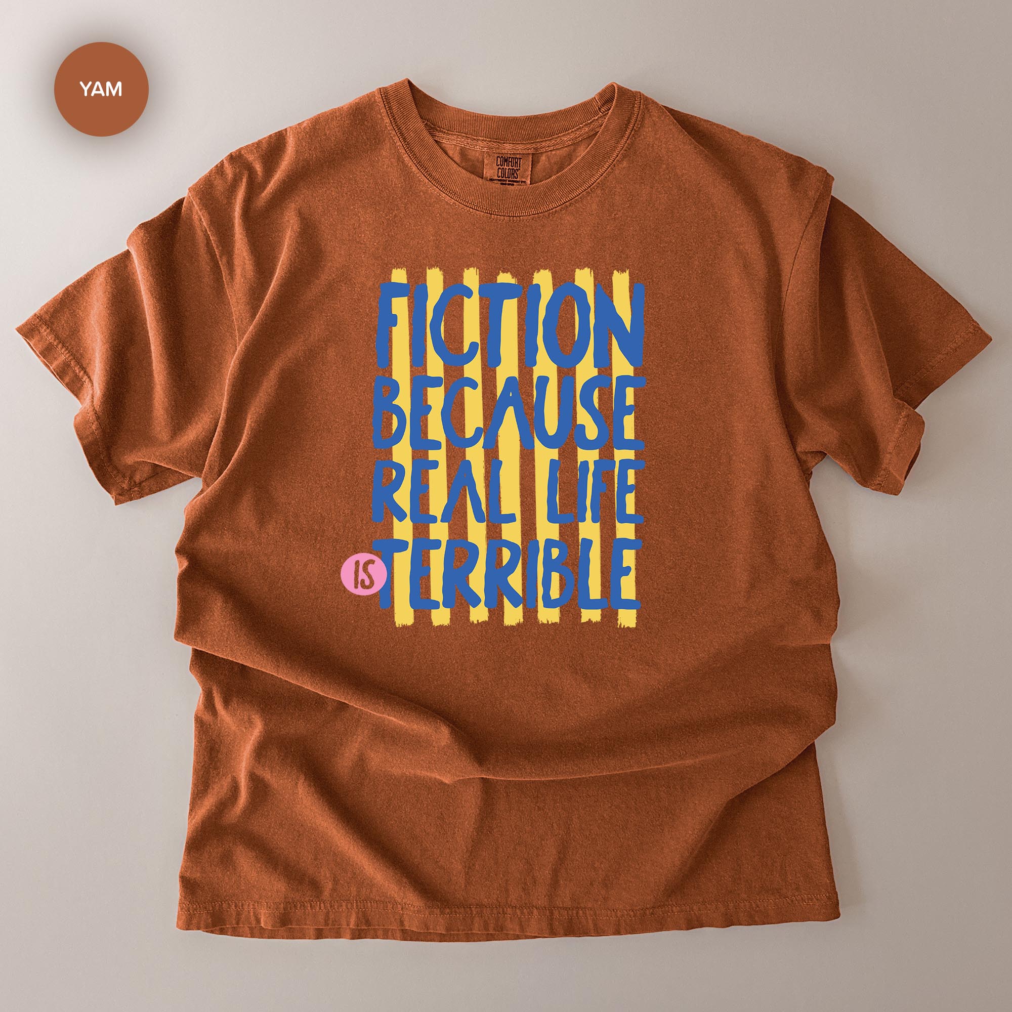 A brown t-shirt with a graphic design featuring the text "FICTION BECAUSE REAL LIFE IS TERRIBLE" in a bold, colorful font.