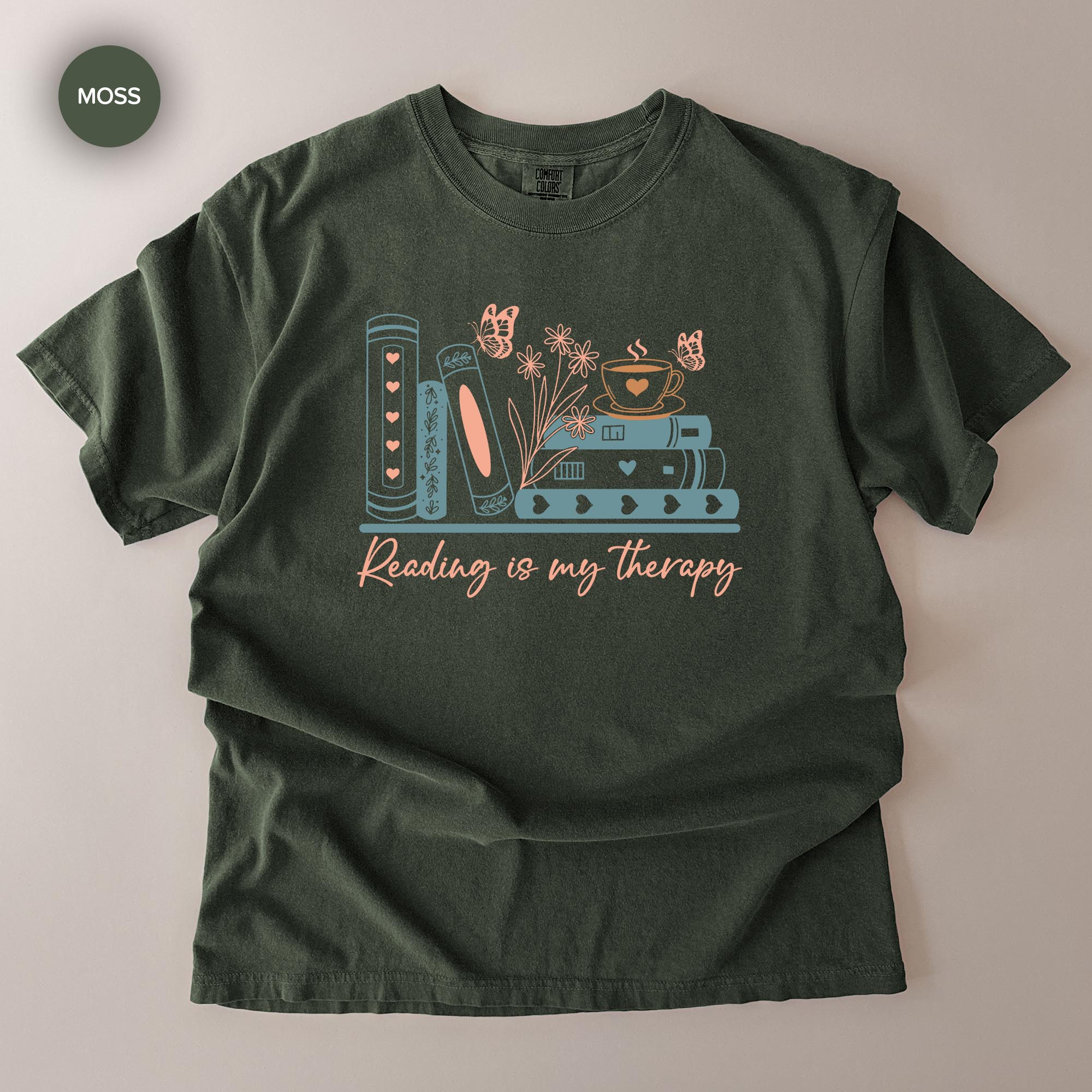 A green t-shirt with a graphic design featuring books, flowers, butterflies, and the text "Reading is my therapy" printed on it.