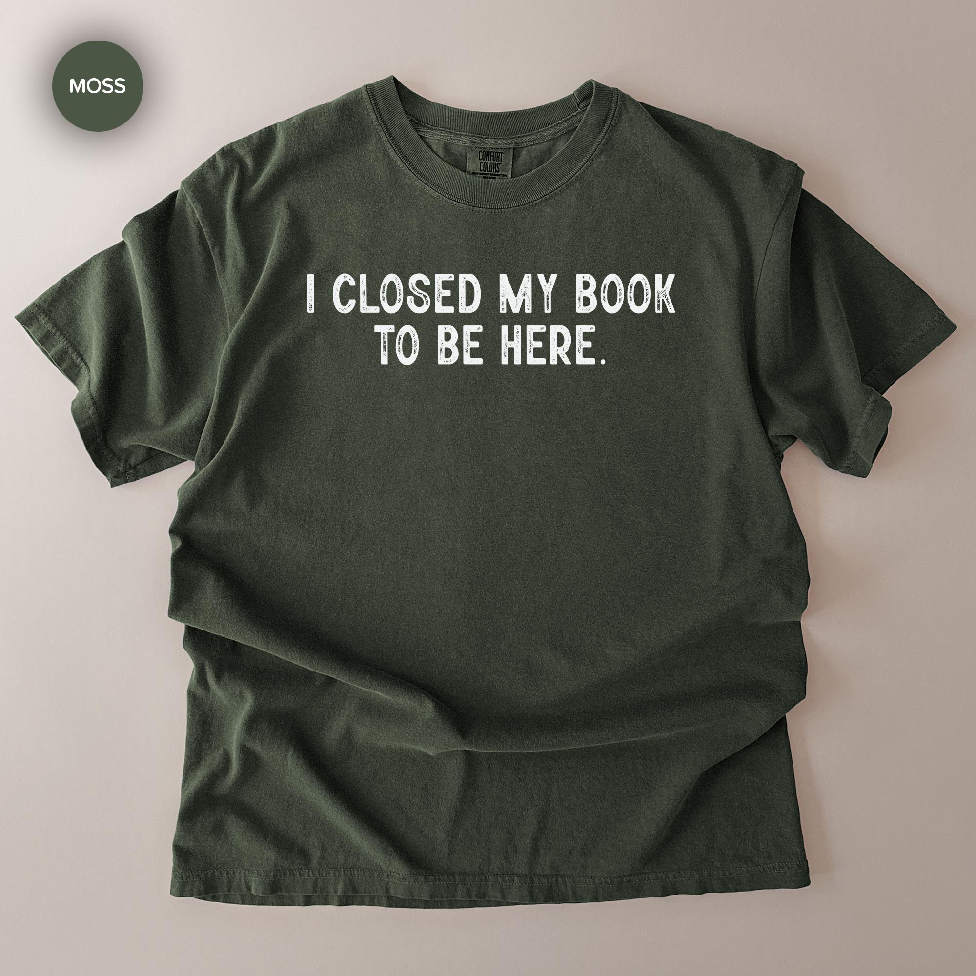 A dark green t-shirt with the text "I CLOSED MY BOOK TO BE HERE" printed on it.